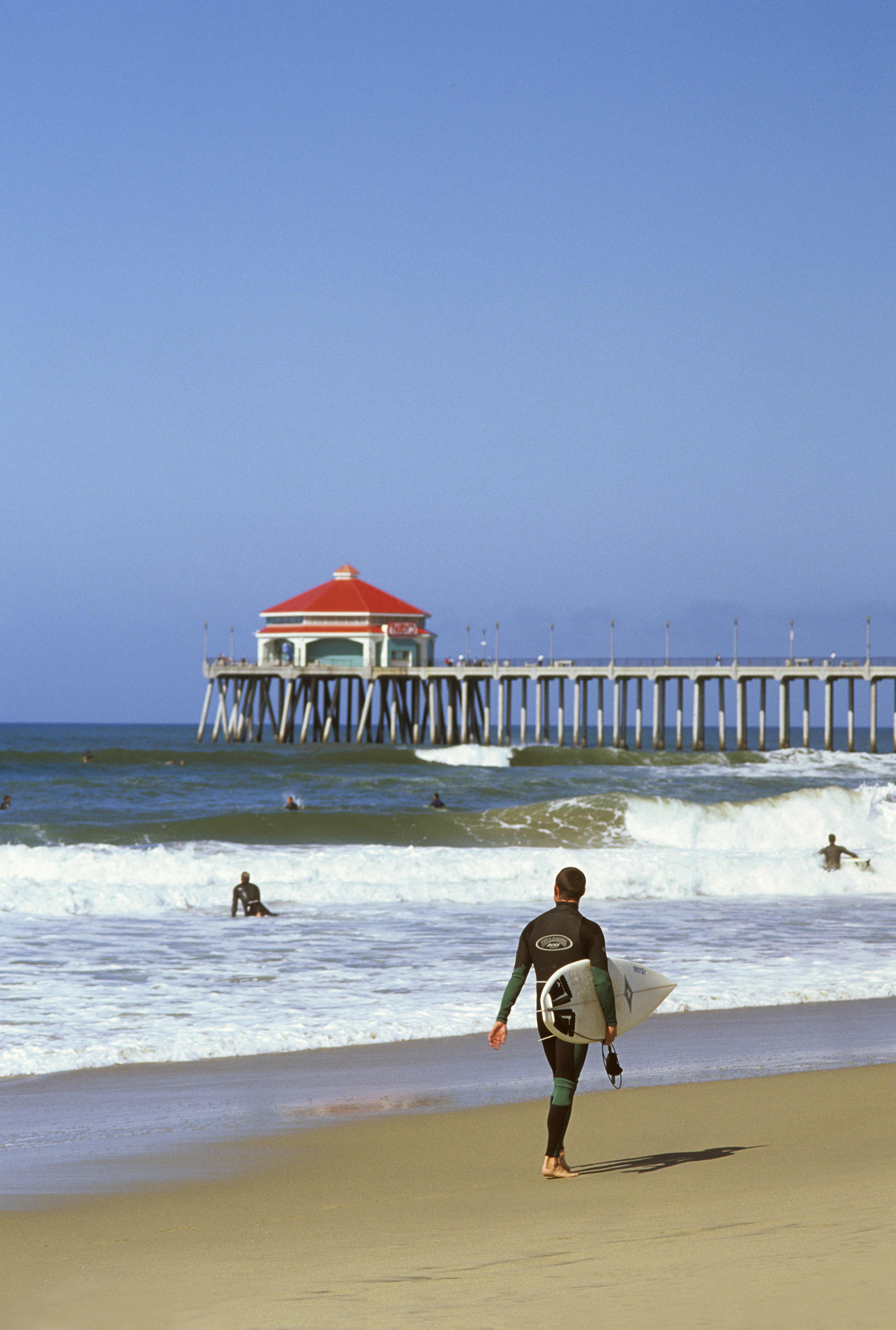 Surfers in black wetsuits hit large white waves against a blue and green ocean with a long pale wooden pier in the background topped with a red-roofed pavilion
