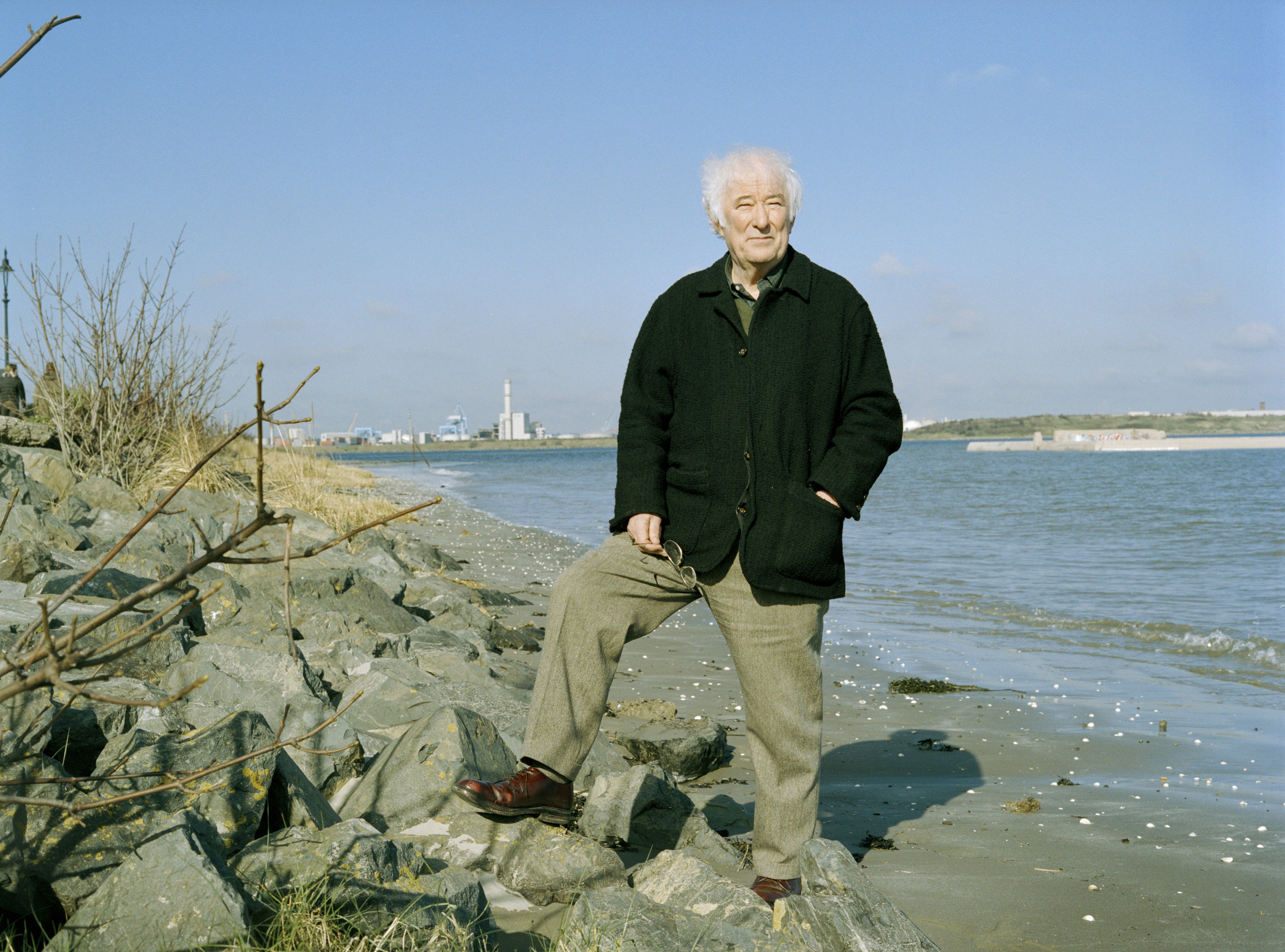 Nobel-prize winning poet Seamus Heaney stands on the Irish coast in tweed slacks, dark brown leather shoes, and a black coat, holding his glasses in his right hand. His left is in his coat pocket. 