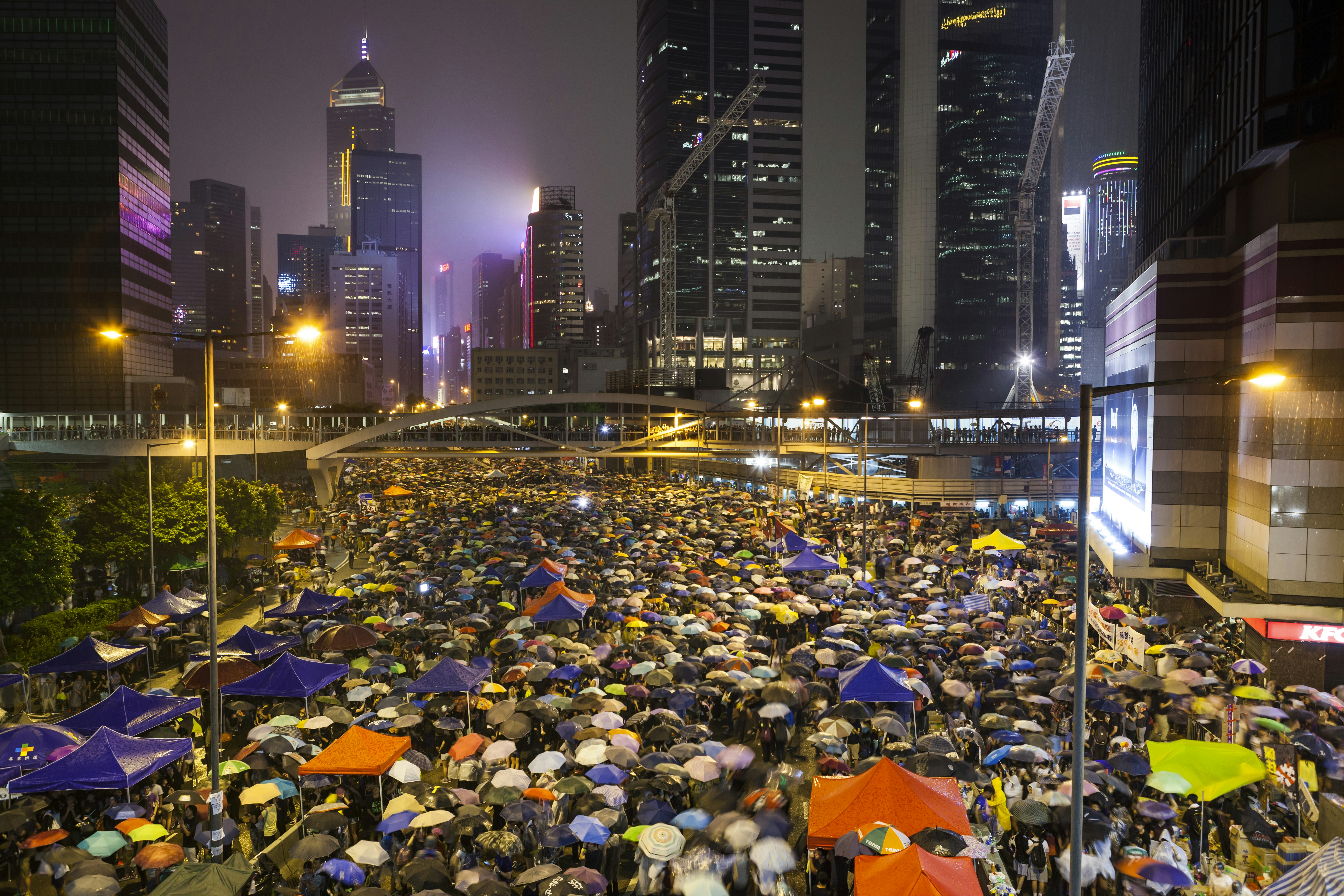 Protestors with umbrellas gather on a street between high buildings at night