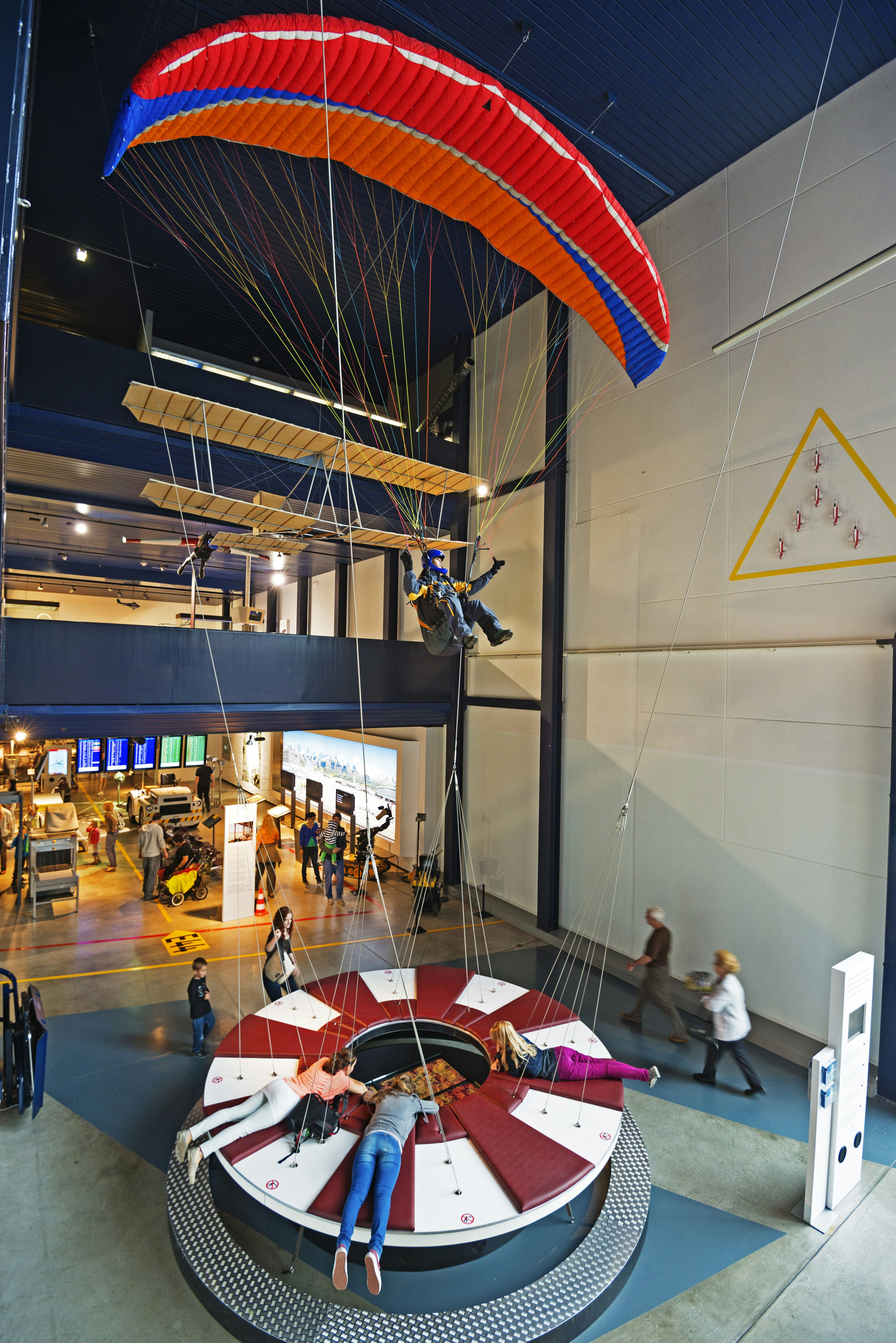 A model of a paraglider with a bright red sail attached with blue tips, hangs from the ceiling of the Transport Museum in Switzerland with a mannequin installed to look like a pilot. Behind, a light wood primitive bi-wing plane also hangs from the ceiling