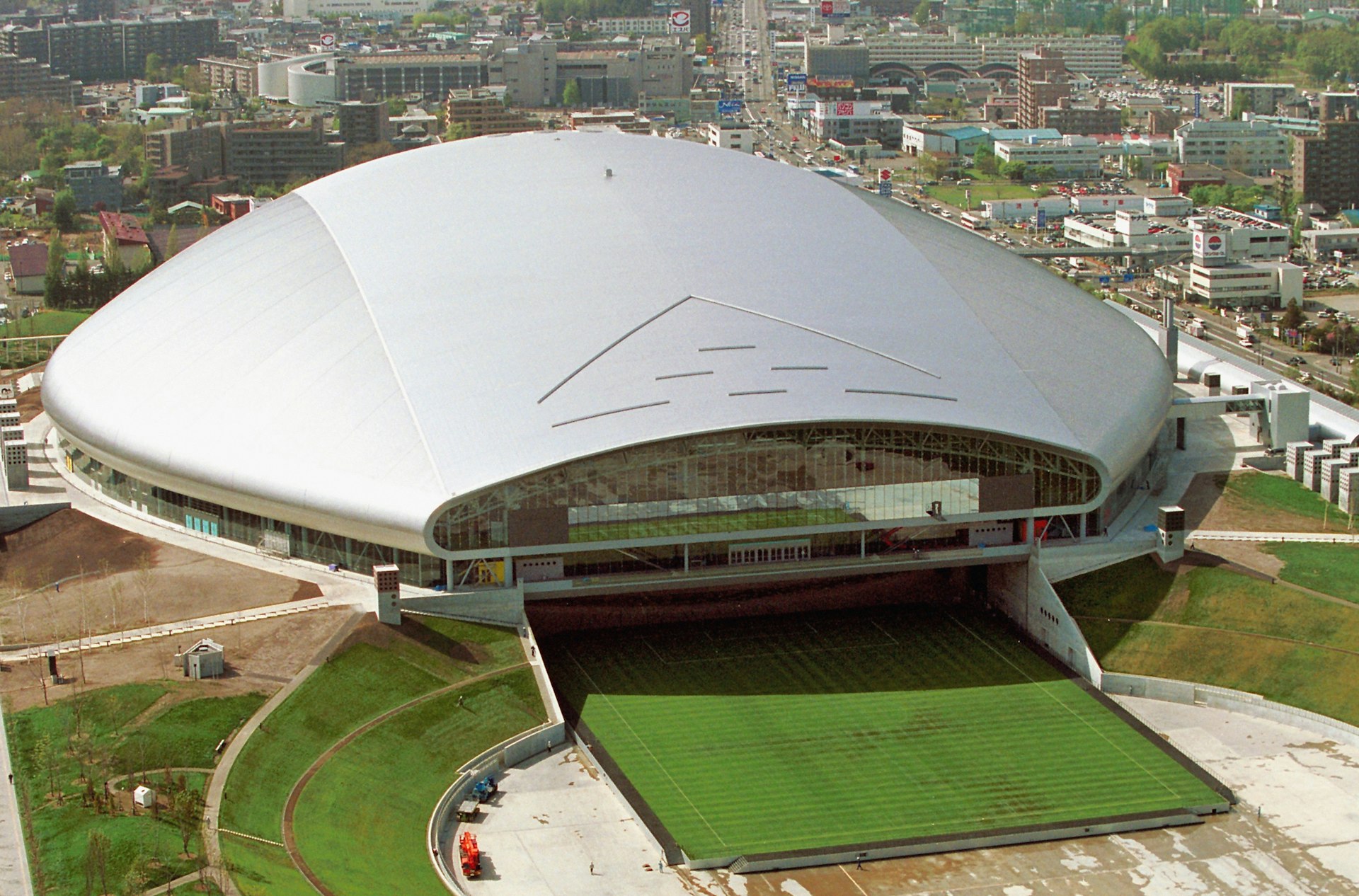 An aerial view of the Sapporo Dome stadium in Sapporo. The stadium is perfectly circular with a sloping domed roof that covers the pitch. City buildings are visible around it.