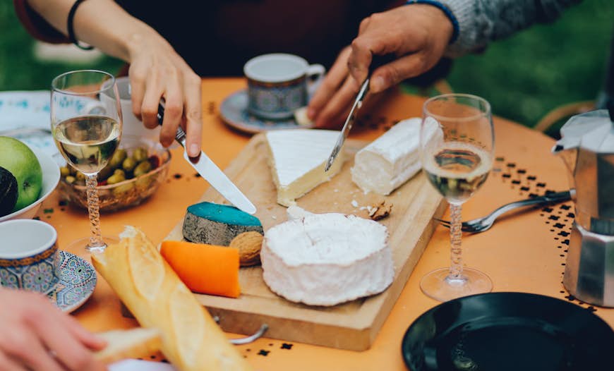 A tabletop showing a selection of bread, cheeses, olives and wine glasses. Diners, sitting around it, are poised with butter knives, ready to dig in.
