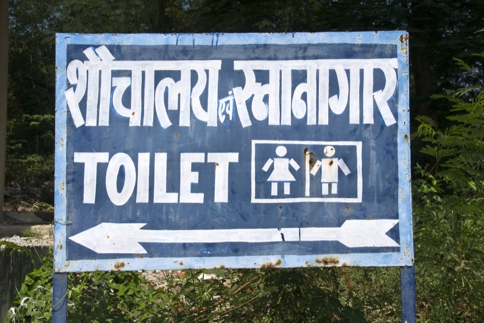 An image of a blue metal sign hand painted with white English and Hindi letters and an arrow pointing to the toilet with symbols for men and women