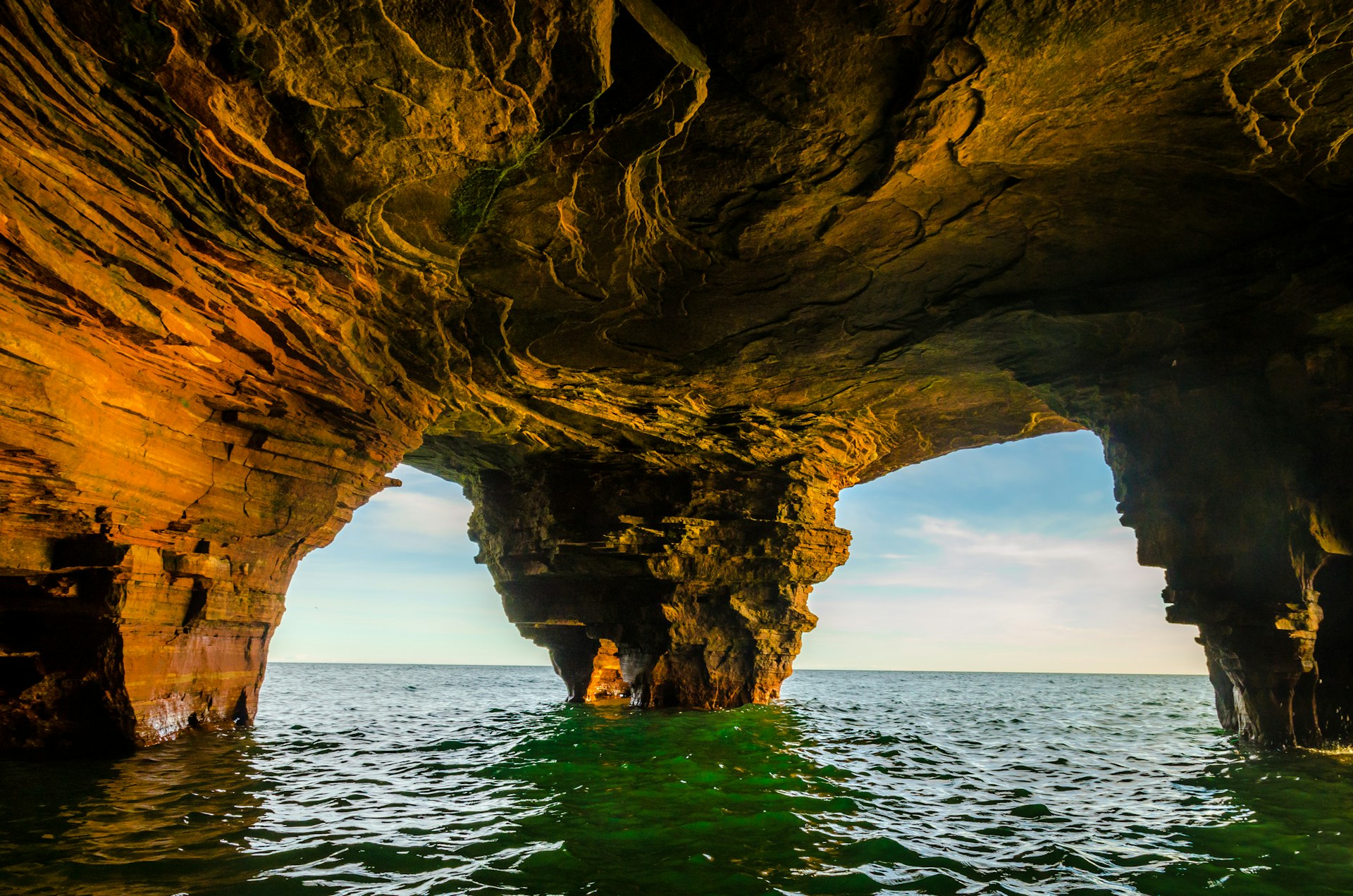 A sea cave near Devil's Island in the Apostle Islands National Lakeshore. The roof of the cave is supported by three thick columns of layered stone, which is illuminated in bright oranges and yellows on the left, dark blacks and yellows in the middle, and deep greenish browns on the right. The light plays on the swirled rock of the cave roof in a way that seems to reflect the deep green waters below. In the far background, a blue sky is streaked with wispy white clouds. Scuba diving in national parks