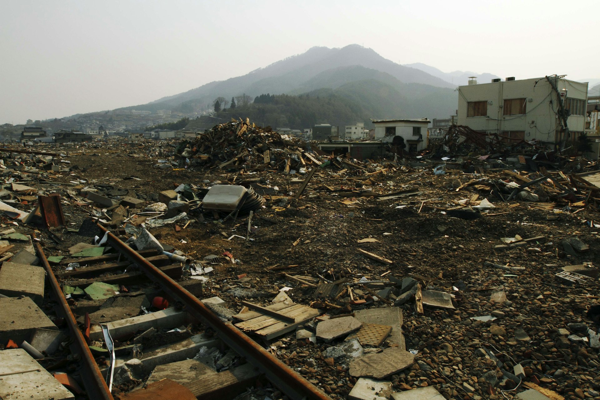 A view of the Japanese city of Ofunato in the aftermath of the 2011 earthquake. Most of the scene is simply rubble, with a train track visible, buried beneath it. A few houses still stand, and in the background, a green mountain peak is visible.