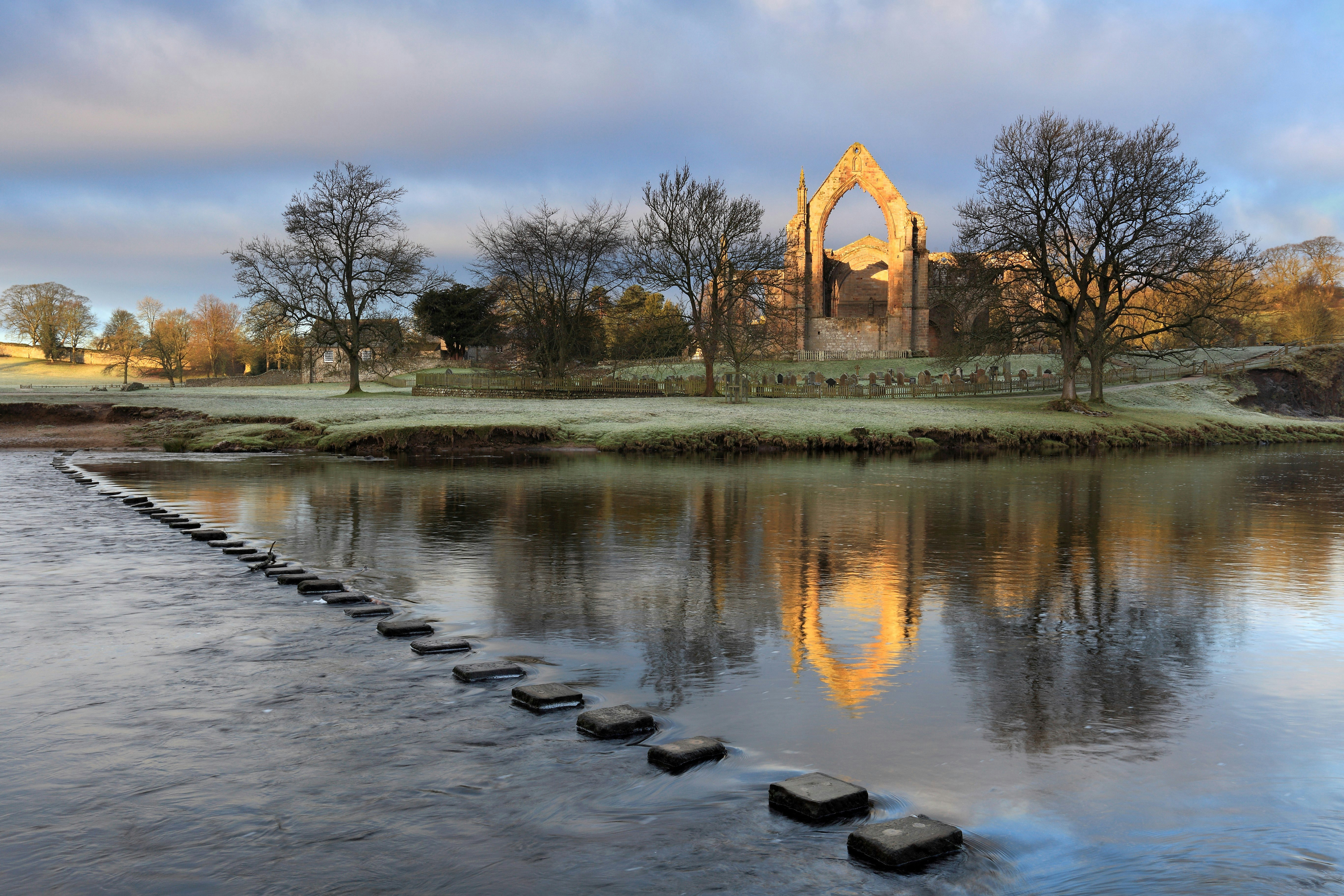 A view of Bolton Abbey, as seen from the opposite bank of the River Wharfe. A series of stepping stones span the river, while the grand abbey is flanked by woodland.