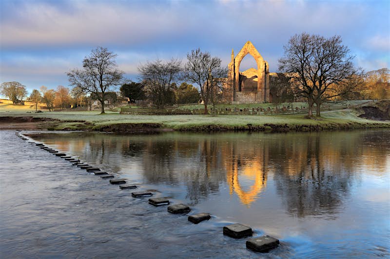 A view of Bolton Abbey, as seen from the opposite bank of the River Wharfe. A series of stepping stones span the river, while the grand abbey is flanked by woodland.