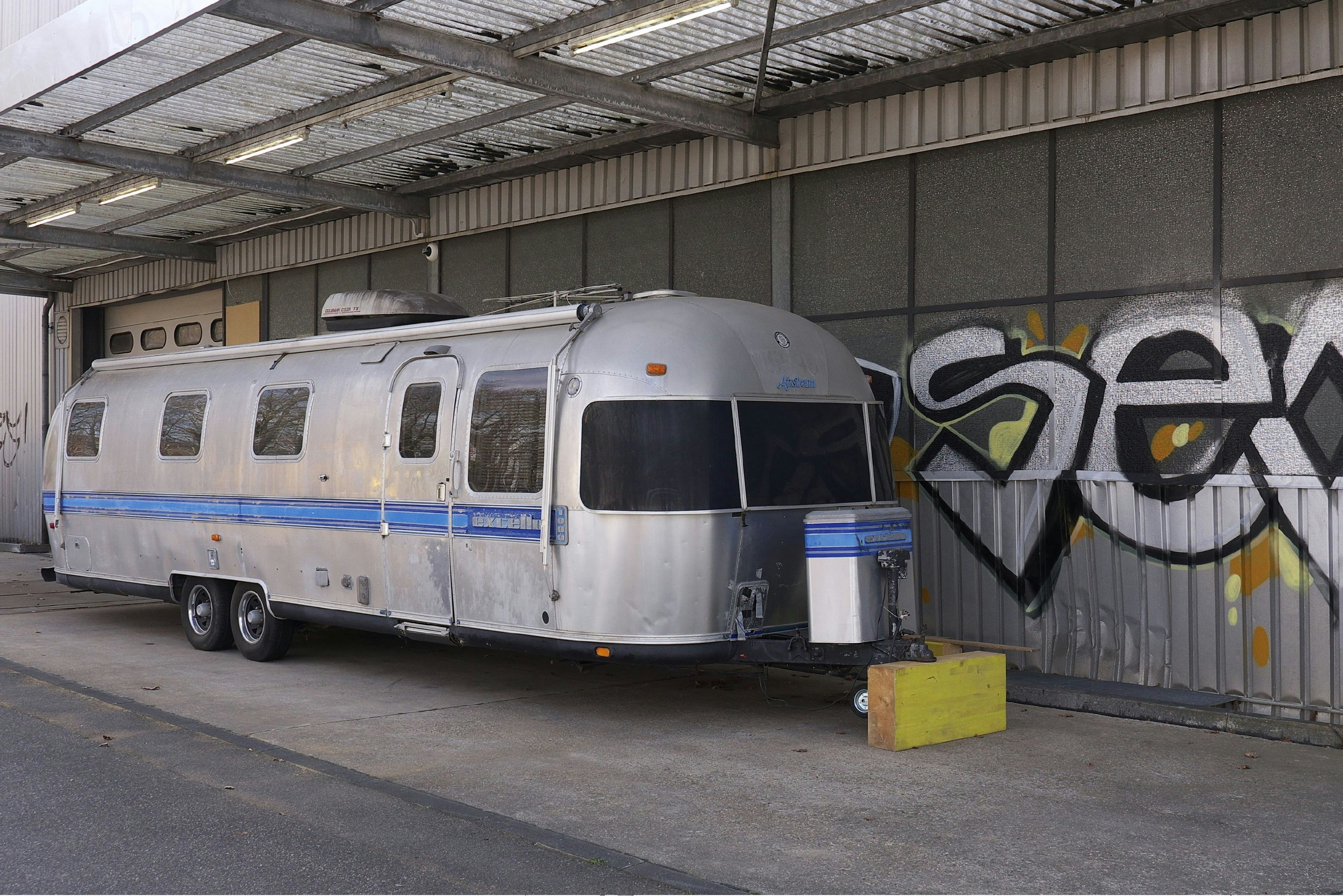 A silver and blue vintage camping trailer sits in front of a concrete wall decorated with graffiti in Bonn, Germany