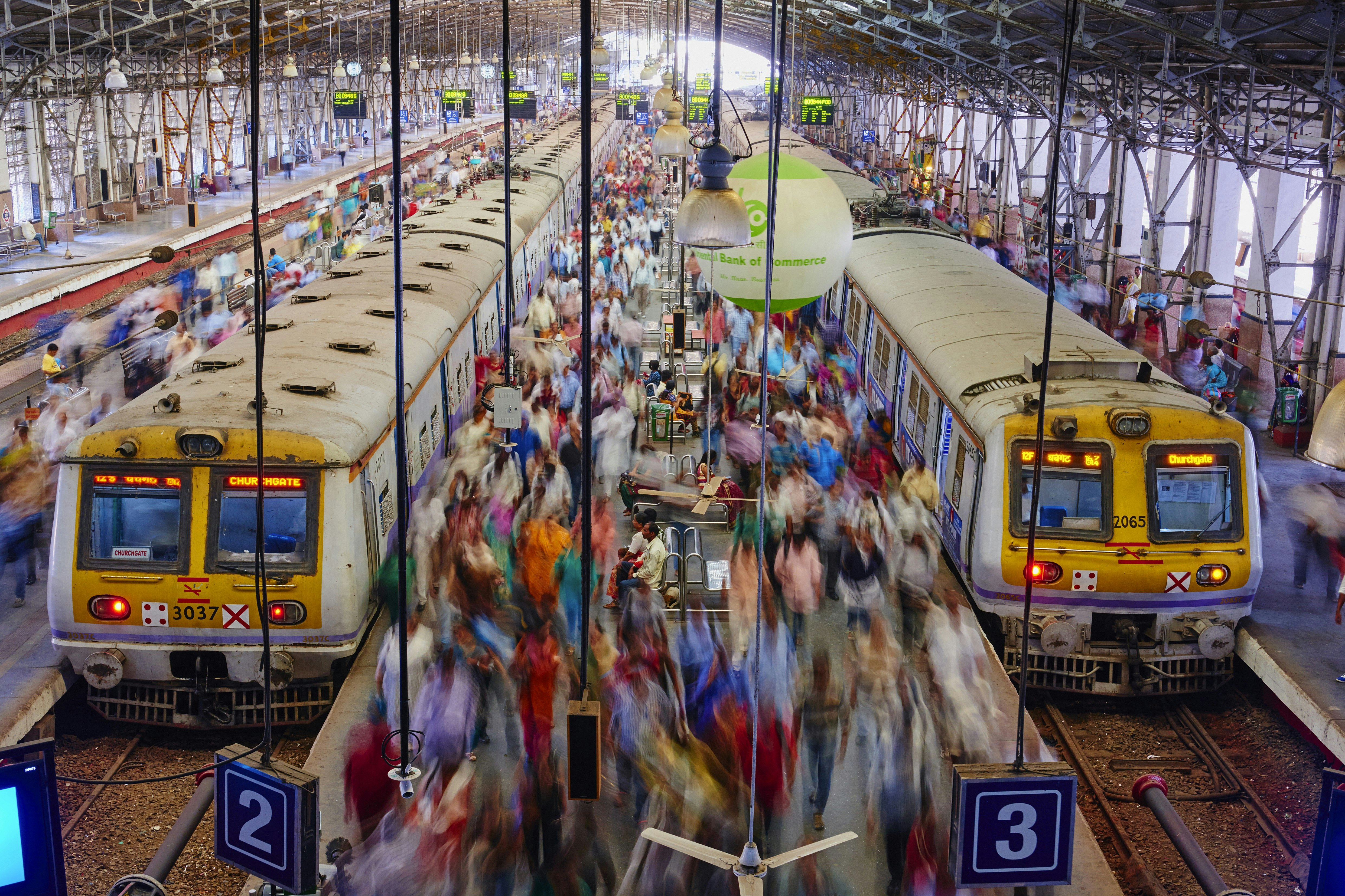 Blurred crowds of people - implying movement - walk along one of the platforms in Chhatrapati Shivaji Terminus, Mumbai. The platform is flanked on either side by trains.
