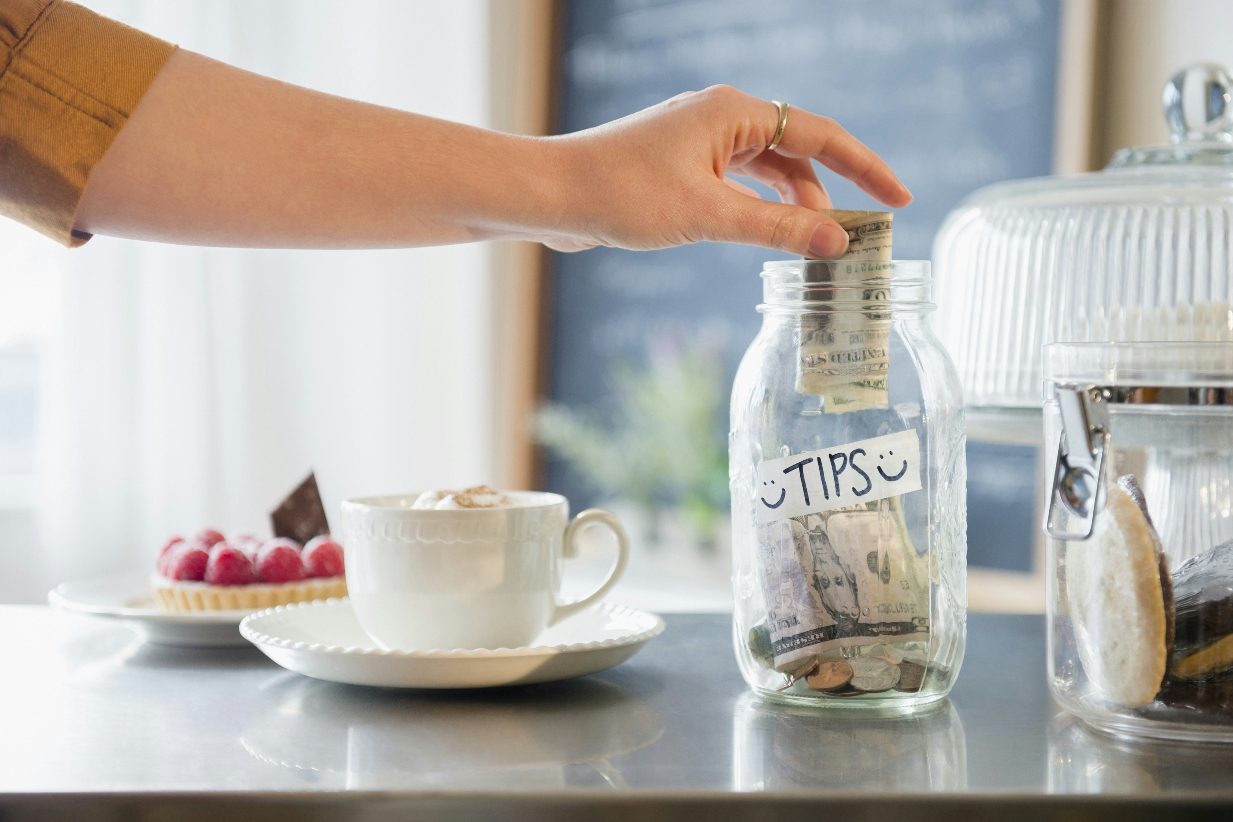 An arm places a dollar bill into a glass jar, which is labelled with a small sign reading 'tips'.
