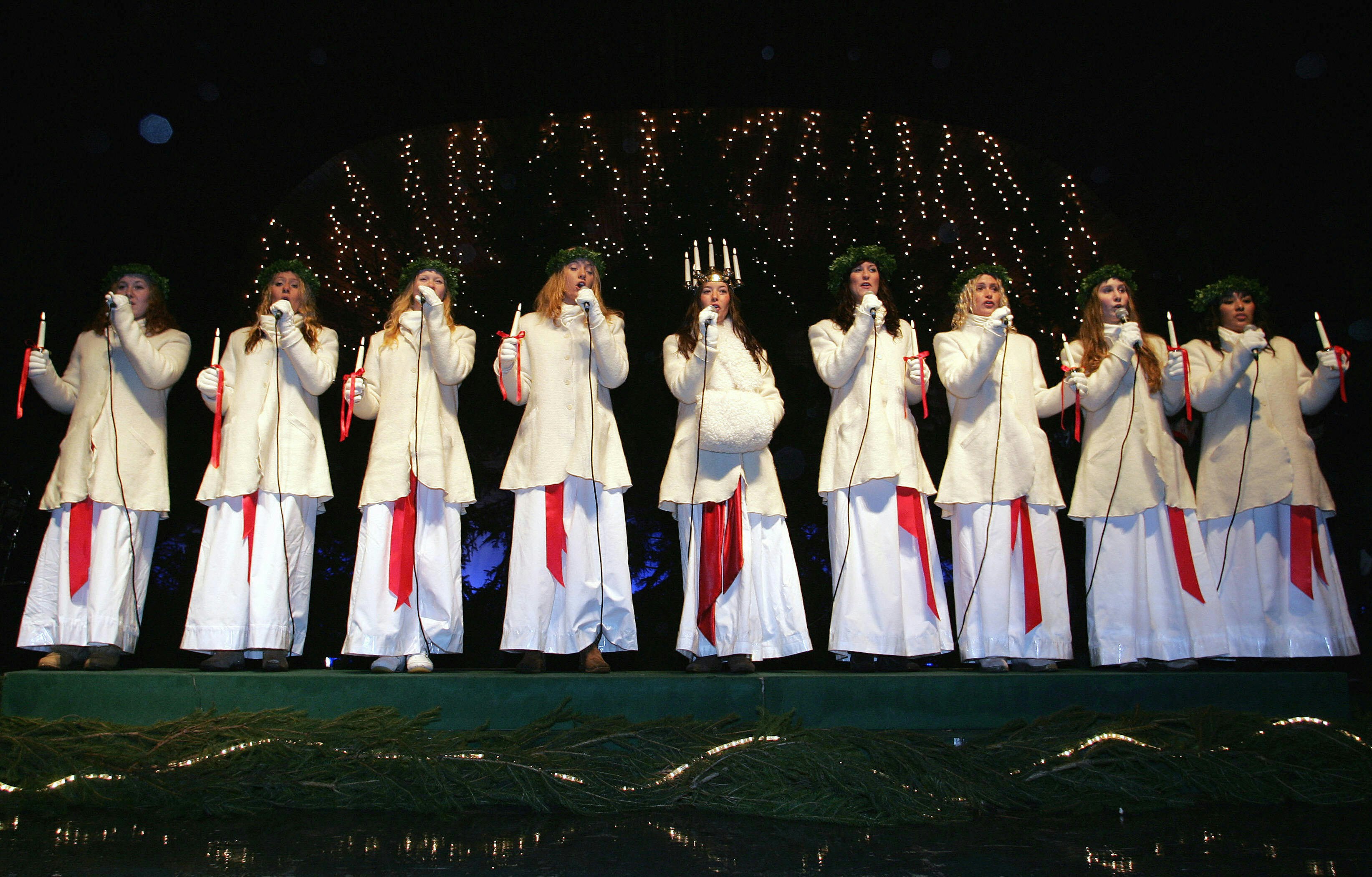 a line of women in white dresses and wreaths on their heads singing on a stage
