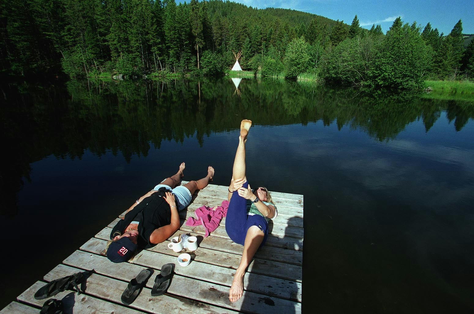 Two women practice yoga on a dock at Feathered Pipe Ranch in Helena Montana. Between them sits a white tea set. In the distance, on the opposite shore of the blue lake, are deep green trees and a white tipi.