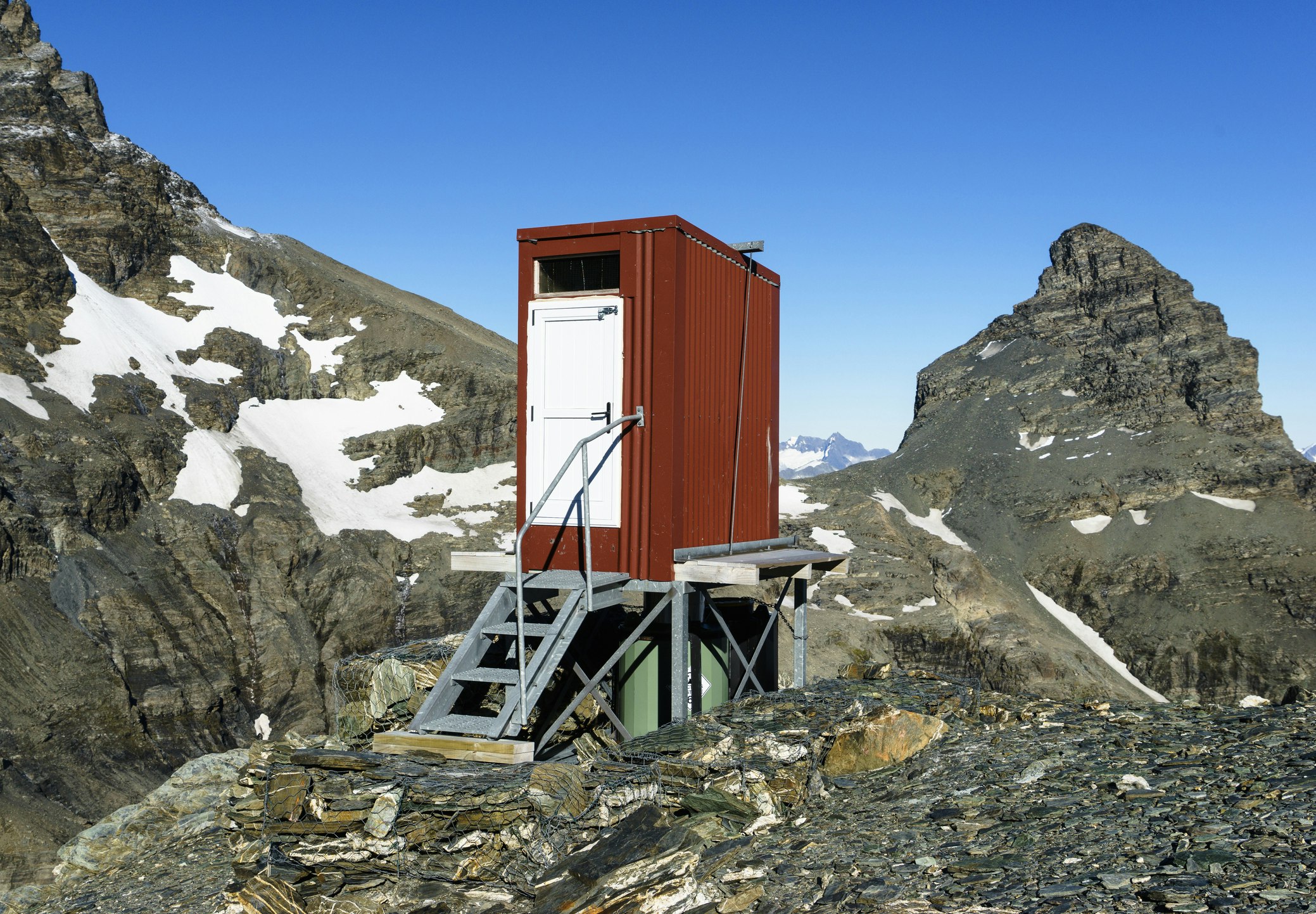 A red corrugated metal shed sits atop a tall, fat green pipe. The front of the toilet shed is bright white, with grey metal steps leading up to it. In stark contrast to the utility of the backcountry bathroom are craggy grey peaks and rock formations, some covered with snow.