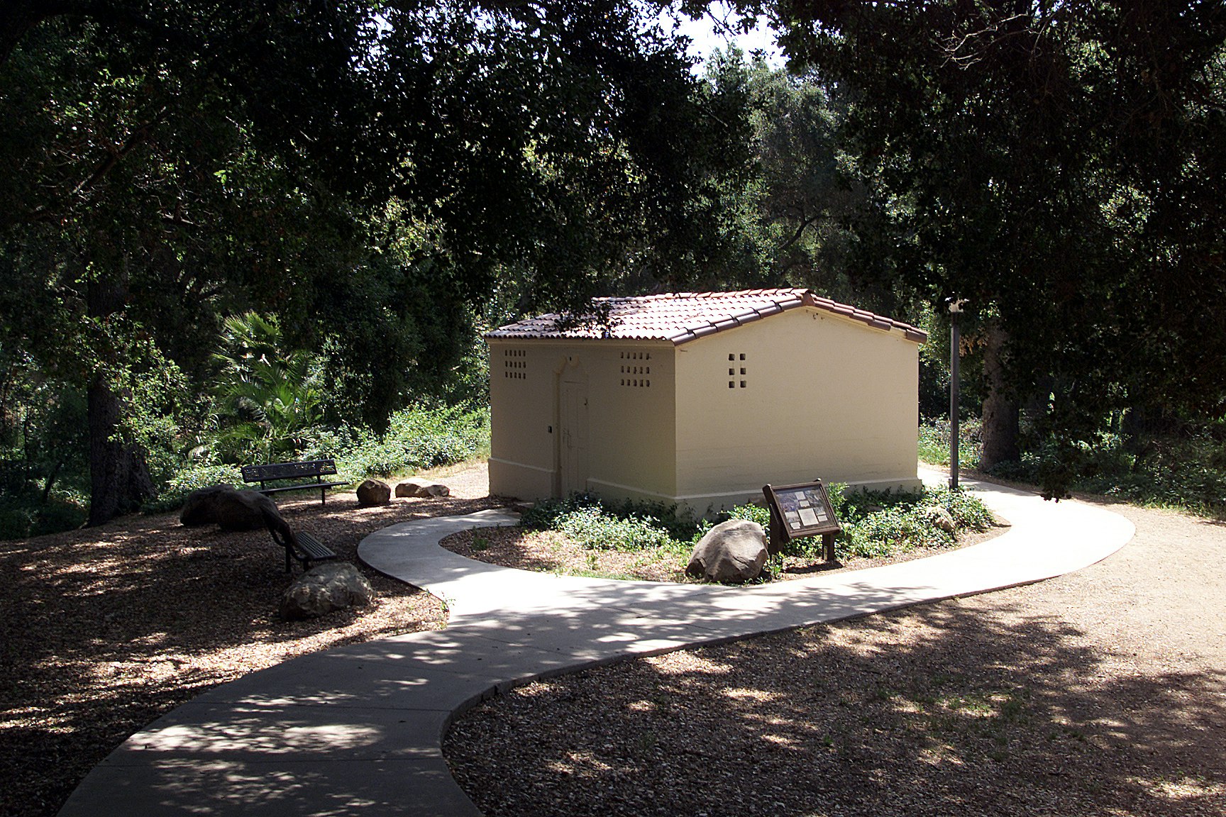 The former Ojai city jail, a small beige square building with tiny square windows and a tile roof its in a grove of trees surrounded by a sidewalk with curving organic forms.