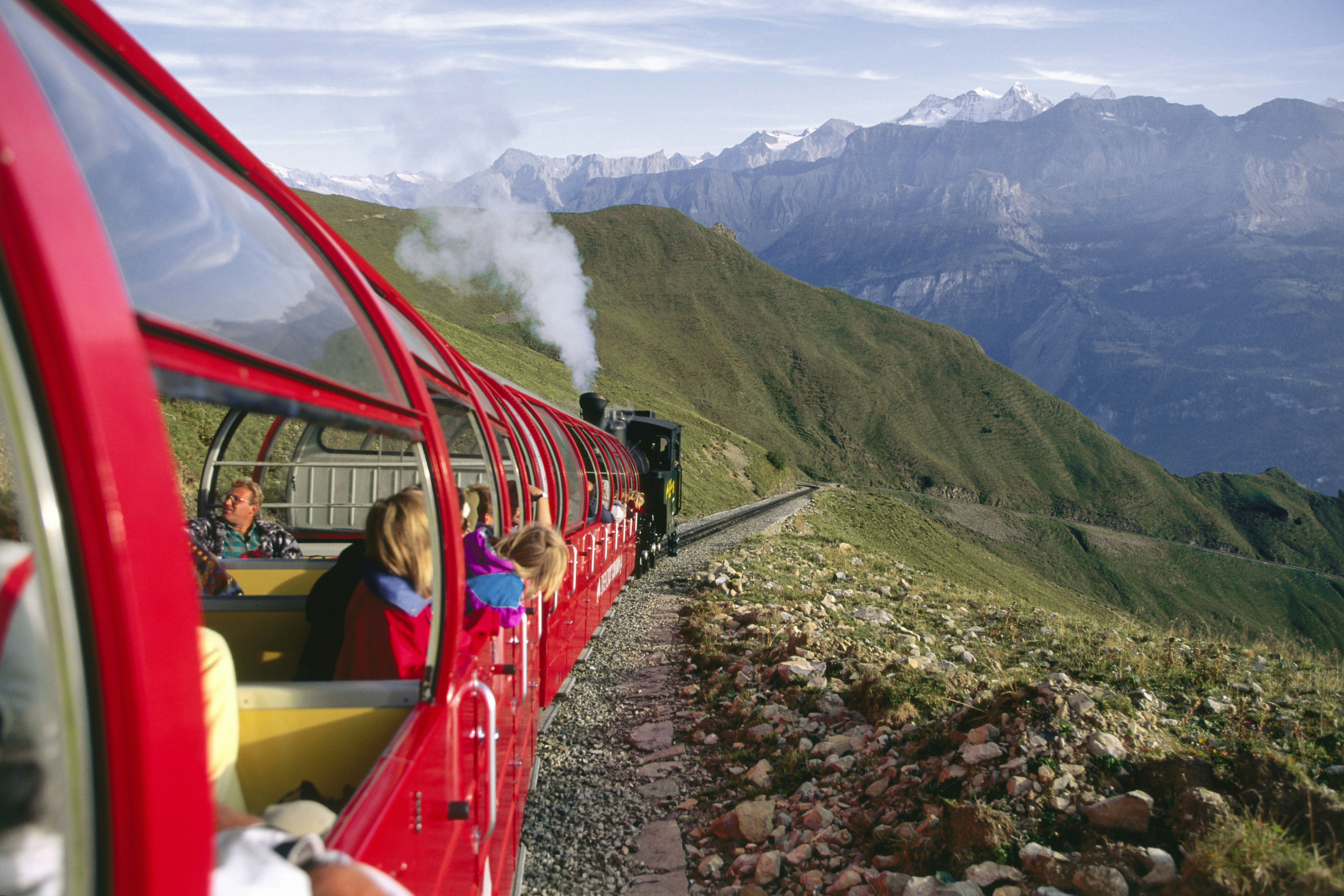 A bright red train with a curved roof made up of as many window panes as possible winds through the Swiss Alps. The photographer is leaning out the window, giving a view back inside of passengers in their seats and one little girl leaning out for a better view of the black steam locomotive pulling the train..