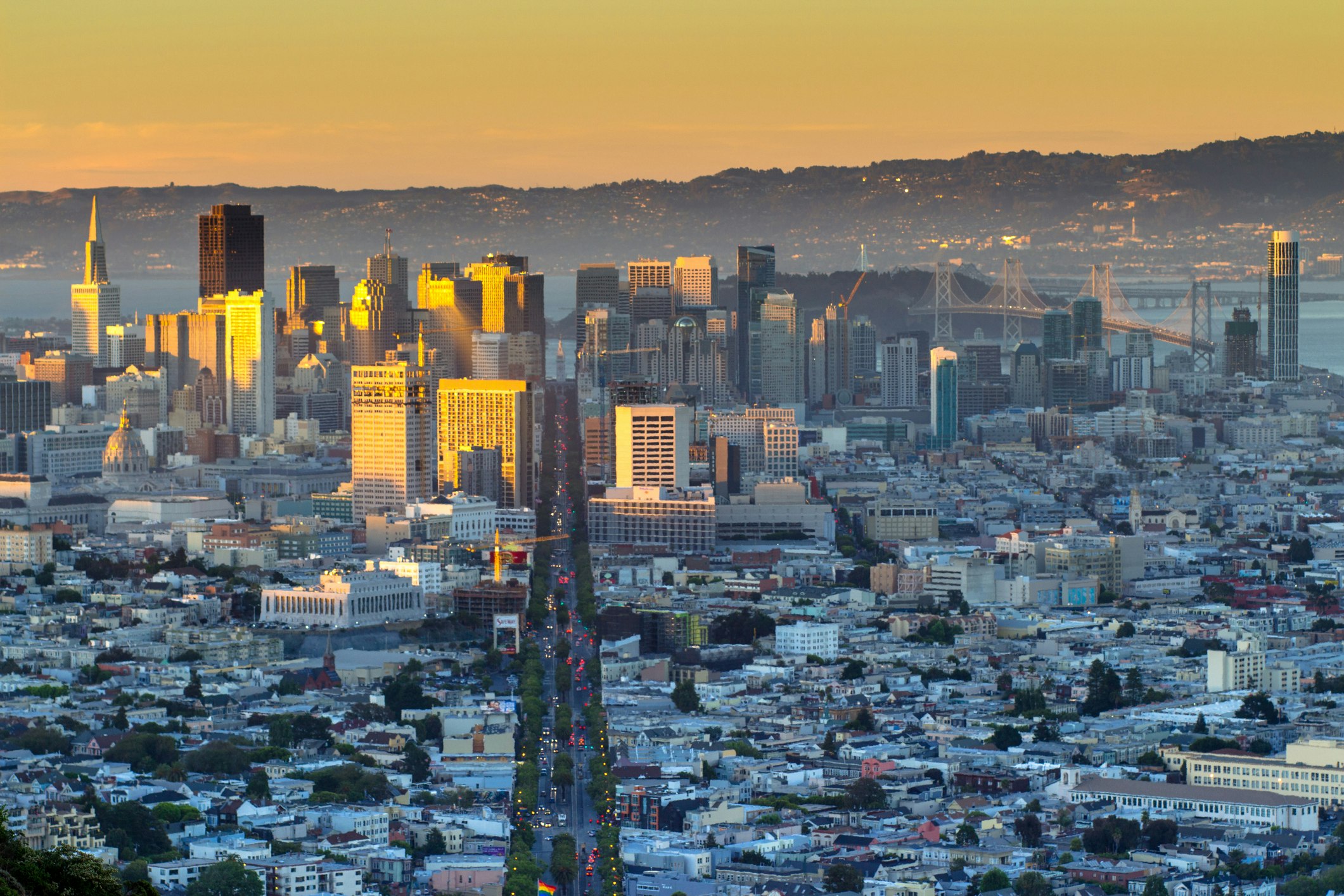 Downtown San Francisco as seen from Twin Peaks. Market Street cuts straight down the center of the frame, and in the background the Berkeley and Oakland hills can be seen across the bay, on the other side of the Bay Bridge.