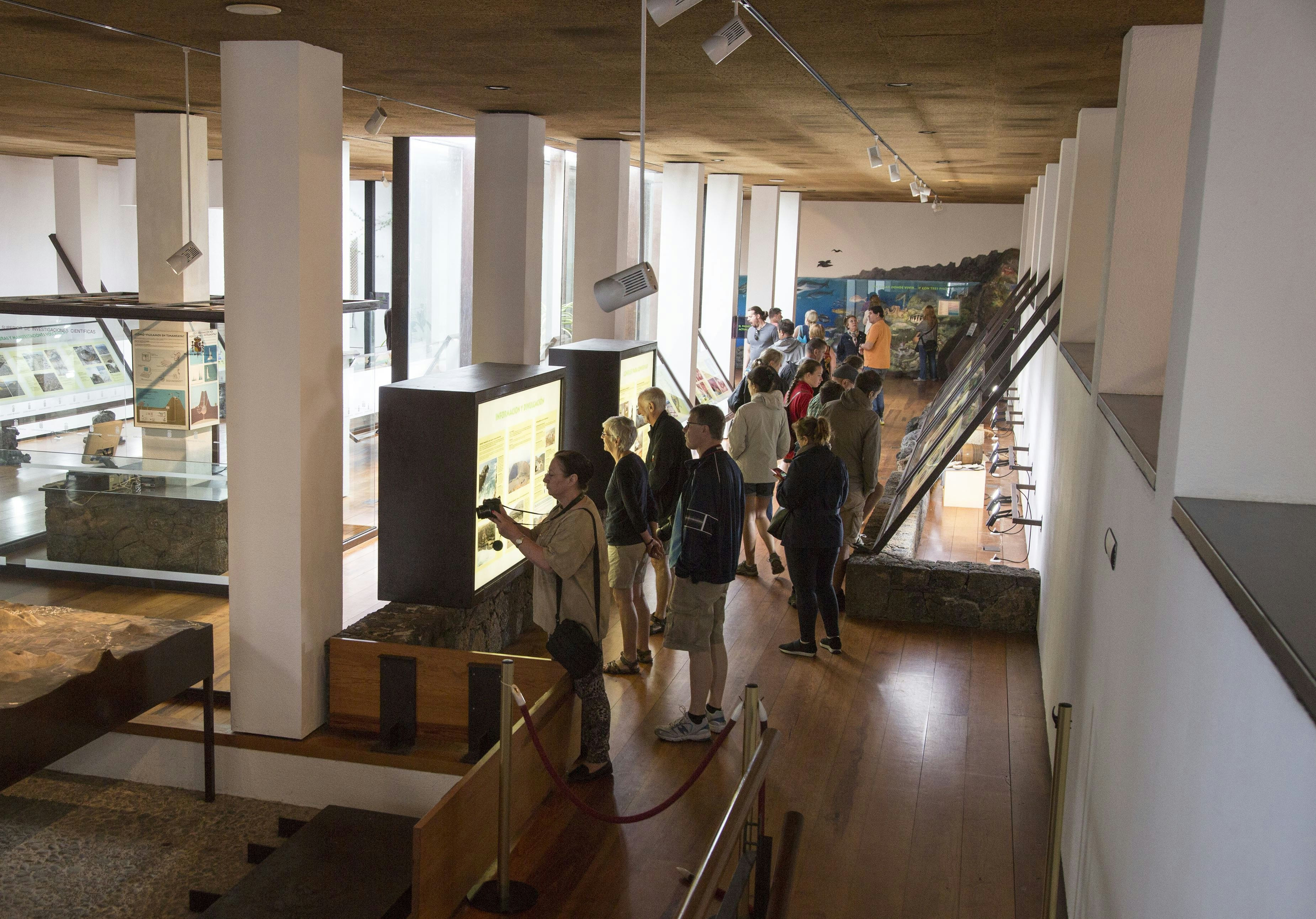 A group of tourists read visual displays and even photograph signage at Timanfaya Volcano Interpretation and Visitors Centre at the Canary Islands in Spain. The room in which the tourists stand is made up of white rectangular columns and smooth white plaster walls with a warm-toned brown ceiling and wooden floor with wide planks. The columns divide the room into two sections full of different exhibits and back-lit informational stations.