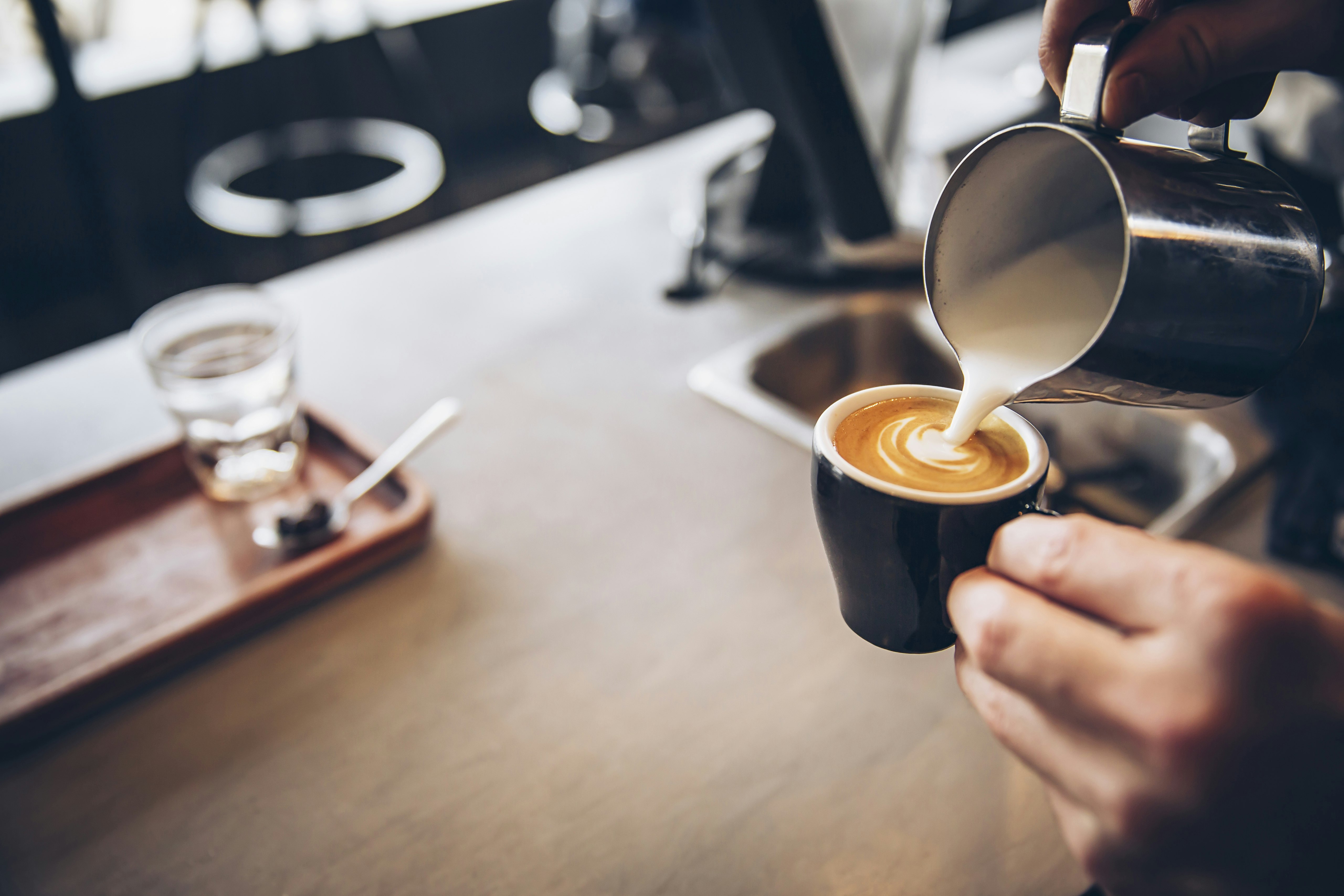 A close-up shot of a barista pouring milk into a small coffee cup over a wooden counter top.