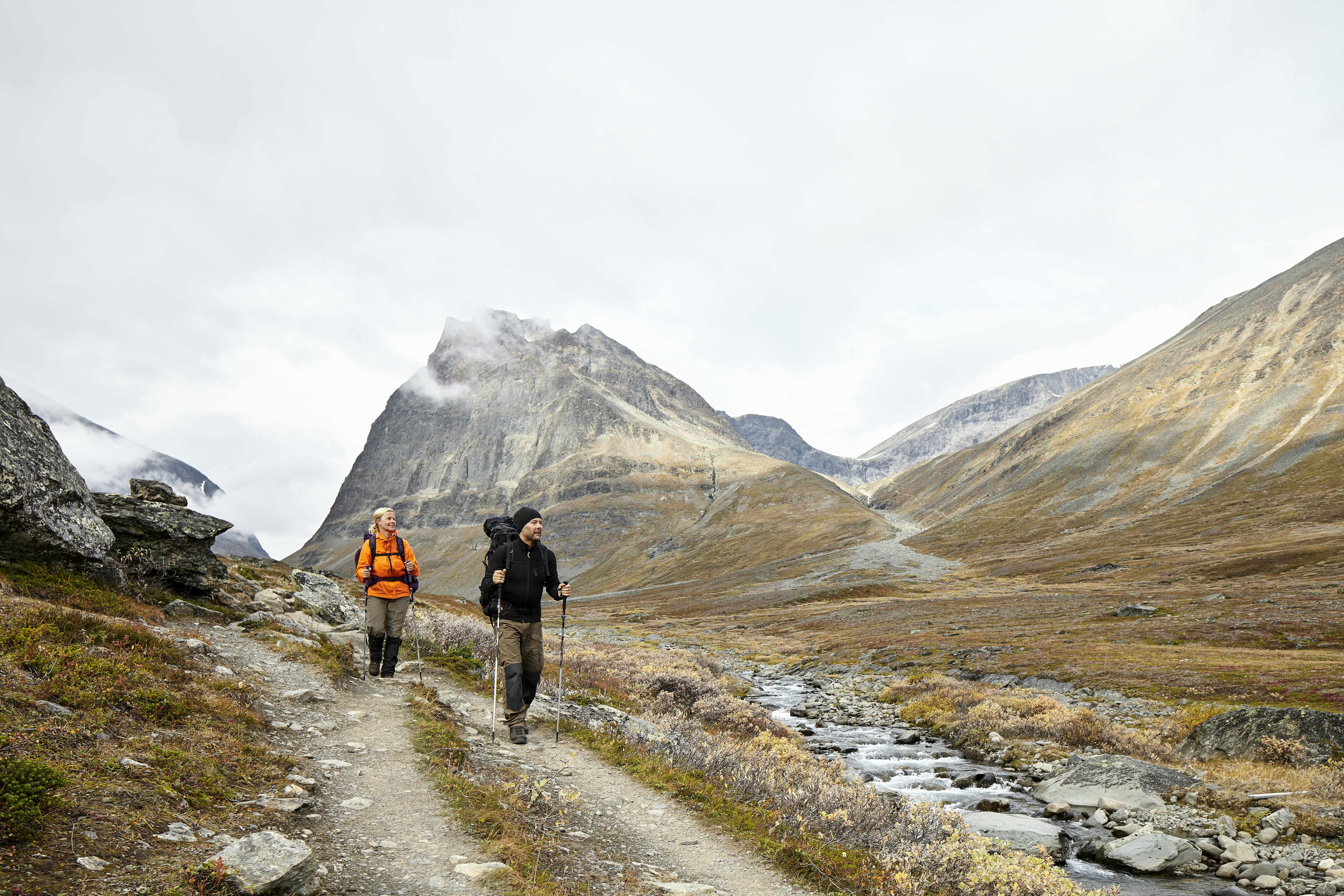 A pair of hikers in dark khaki slacks, one in a black jumper with a black backpack and the other with a bright orange jacket and black pack, both with trekking poles, approach the viewer along a two-track trail between fog-shrouded massifs in Sweden