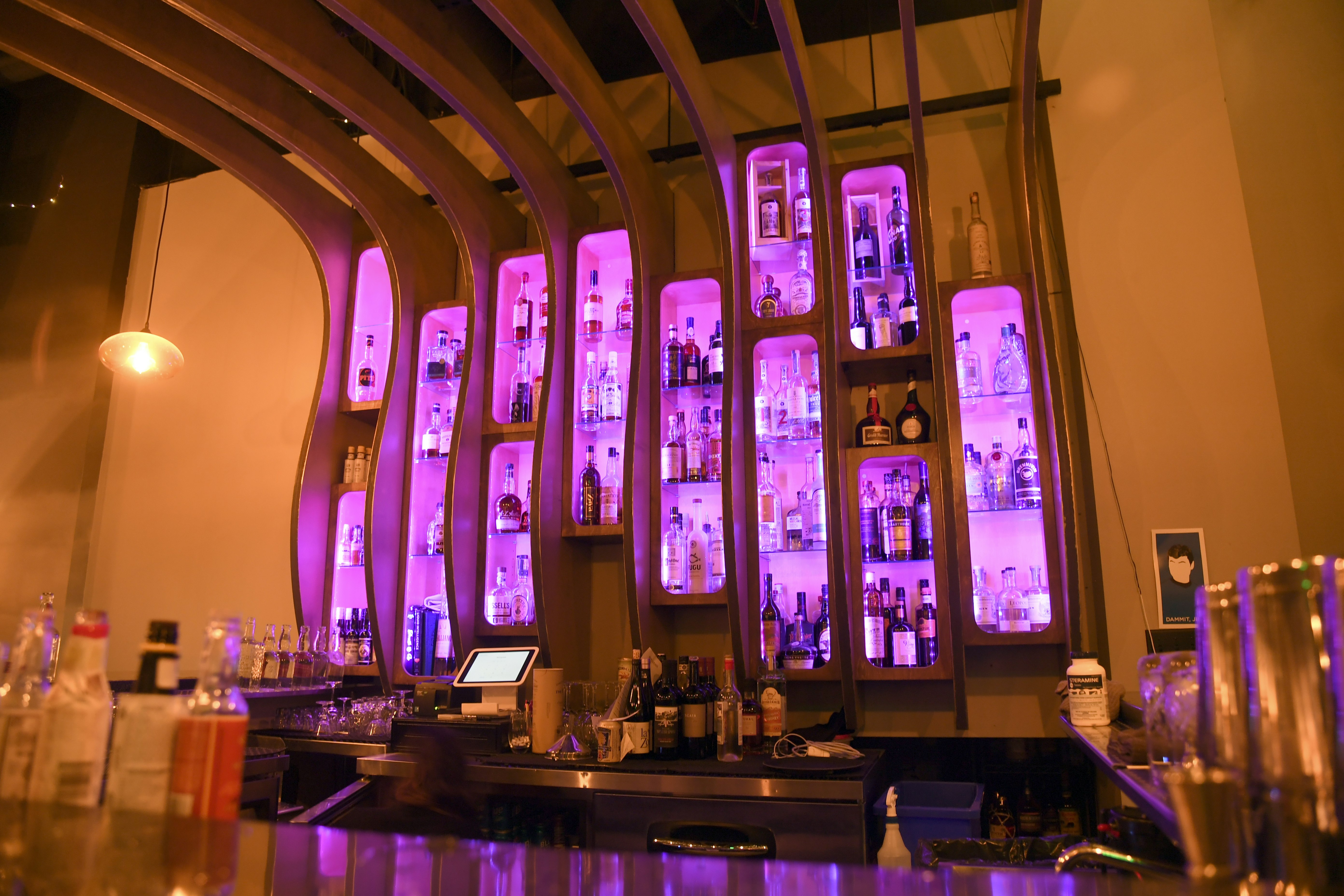 Bottles of liquor are displayed in purple glowing shelves divided by curved wooden ribs, a contemporary take on Midcentury Modern aesthetic forms