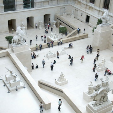 Aerial view of the Cour Marly, a courtyard in the Louvre filled with sculptures from the 17th and 18th centuries. The white courtyard is filled with visitors looking at the exhibits.