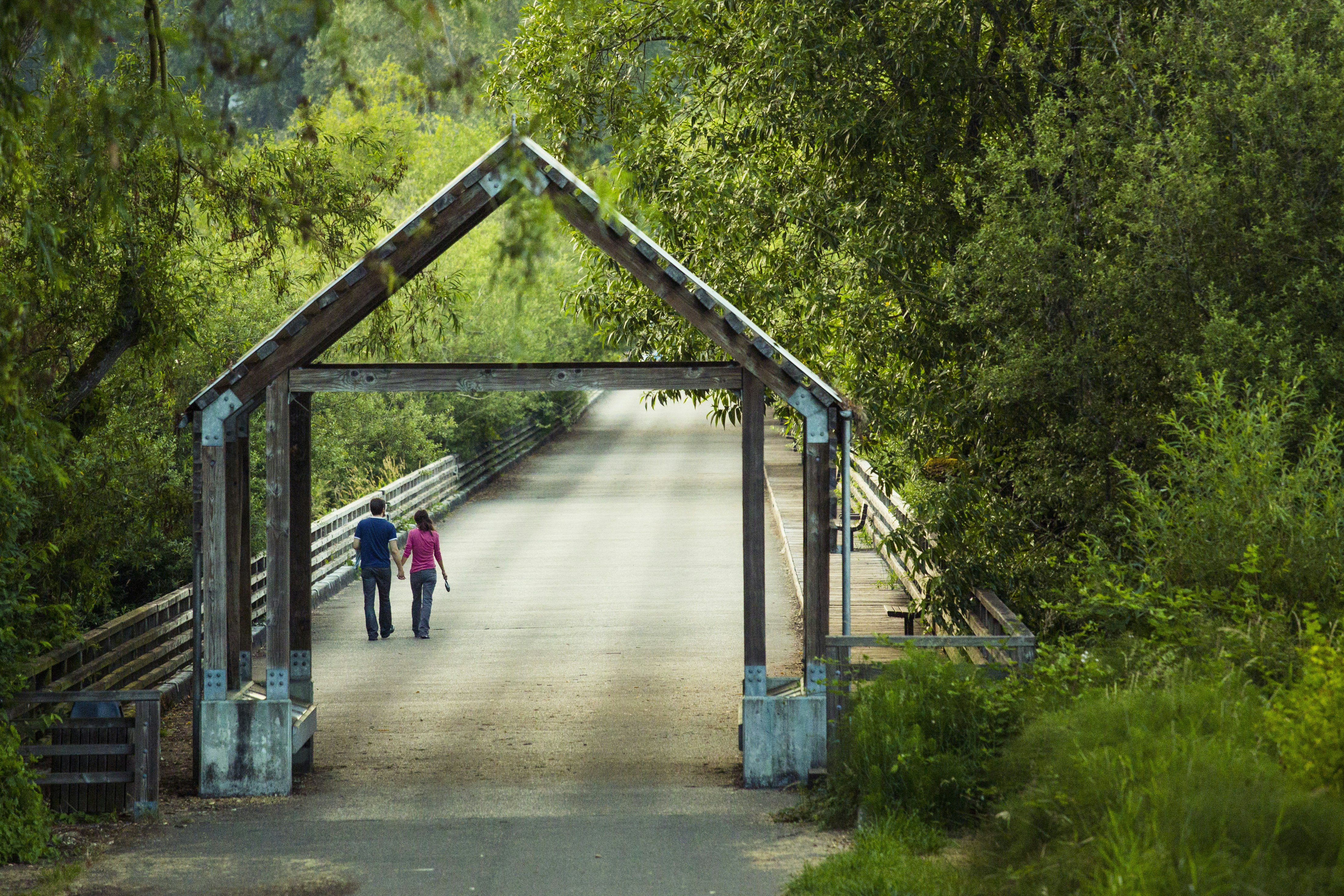 A man in a blue t-shirt and jeans and a woman in a pink t-shirt and jeans walk hand and hand down a paved trail in Juanita Bay Park in Kirkland, Washington, surrounded by green trees on either side of a long fence of wooden rails. In the immediate forground is a gate made of wood beams and metal joists in the outline of a house like a child might draw, with square walls and a triangle roof