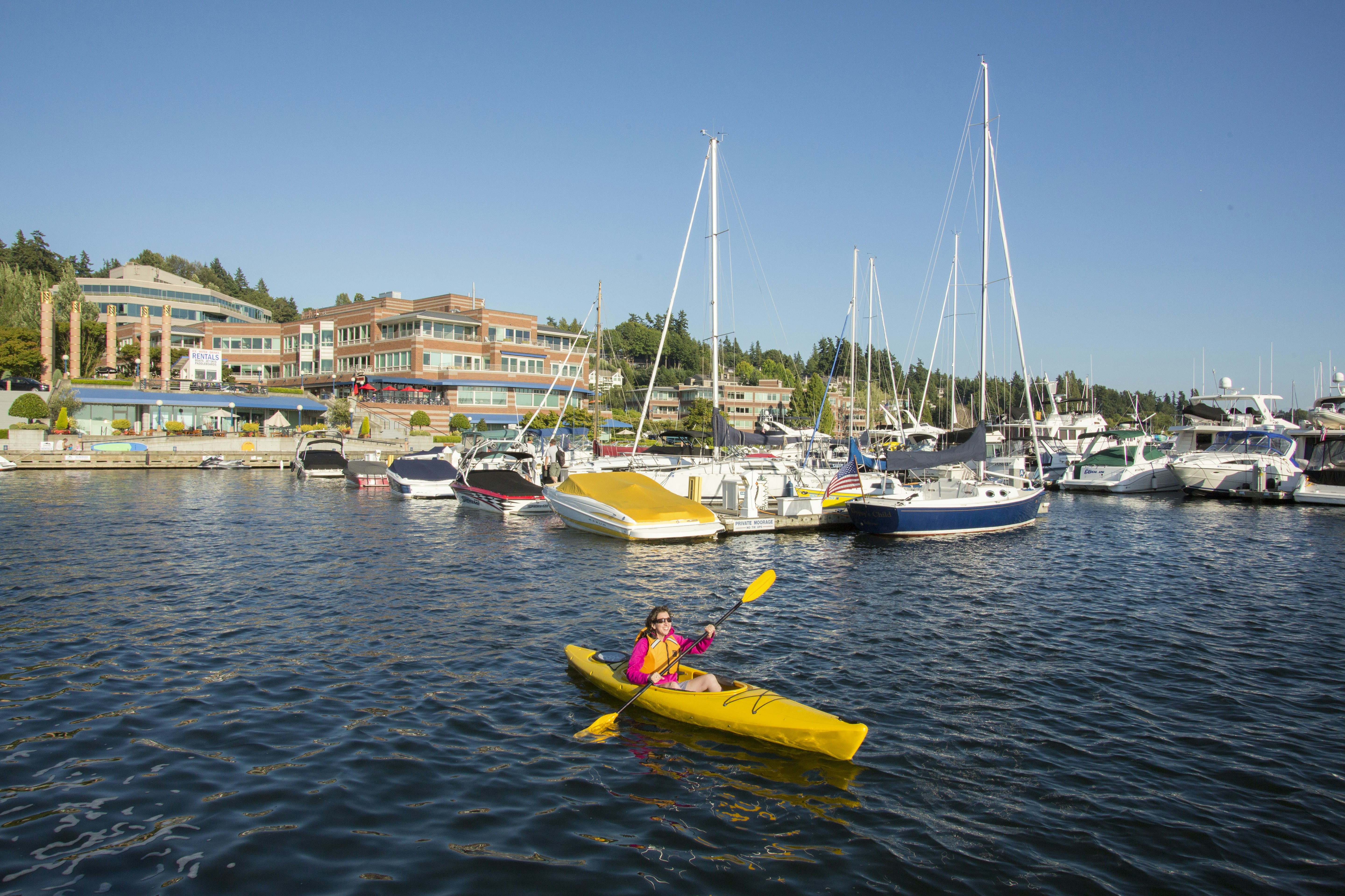 A woman in a hot pink long-sleeved top and a yellow life preserver paddles a yellow kayak with a yellow and black paddle in deep blue waters in a Kirkland, Washington marina full of sailboats, house boats, and a large brick building on a hill