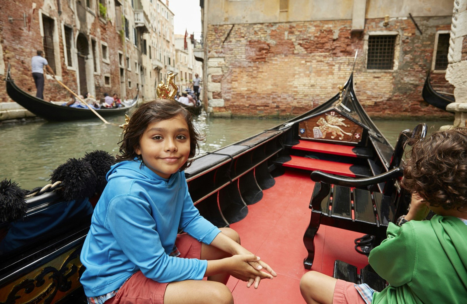 Two boys sit on a red gondola floating through Venice, one boy is looking at the camera and the other is looking away