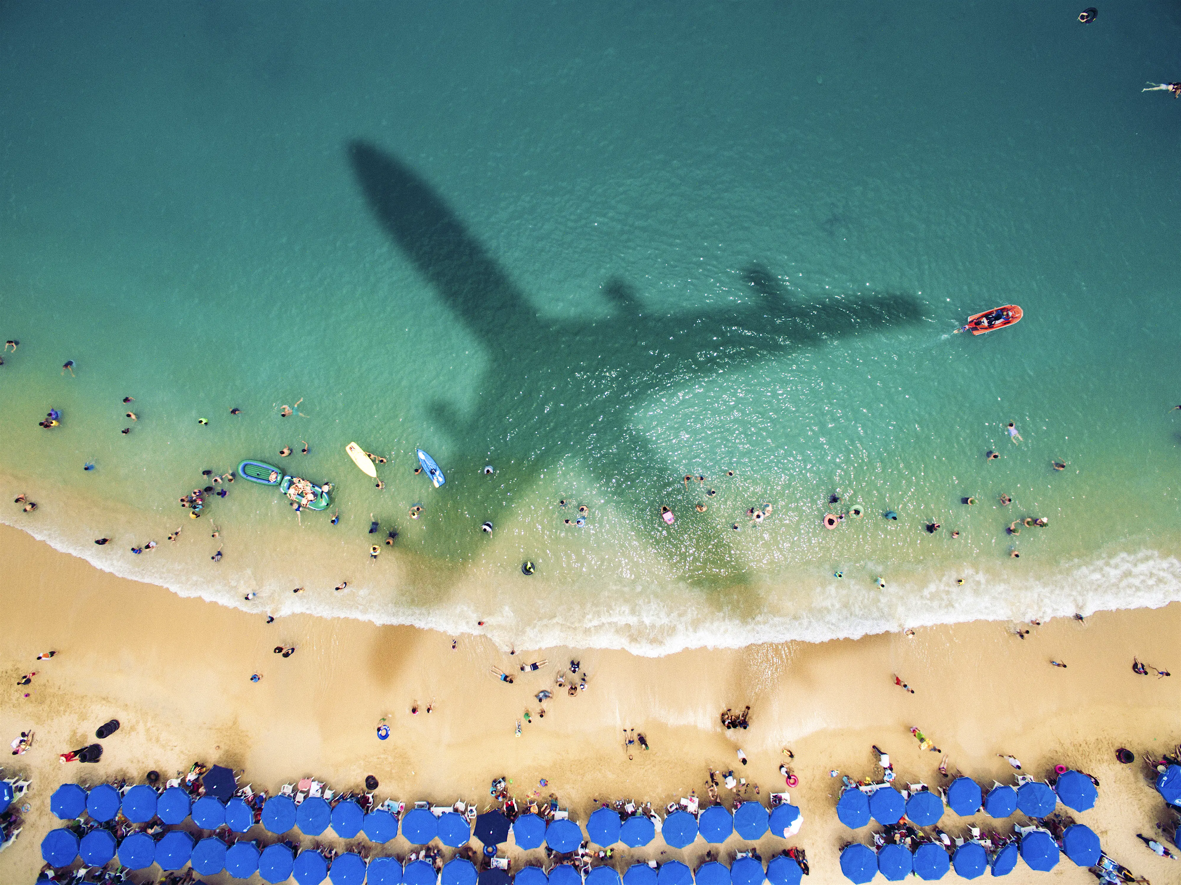 carbon-offsets-holiday-travel-beach.jpg The shadow of a large jet plane falls over a beach with picturesque white sand, deep turquoise waters, and two long, waving rows of blue beach umbrellas. In the white surf break are the tiny dots of swimmers and beachgoers, as well as a red boat and a couple inflatable beach toys and two paddle boards.