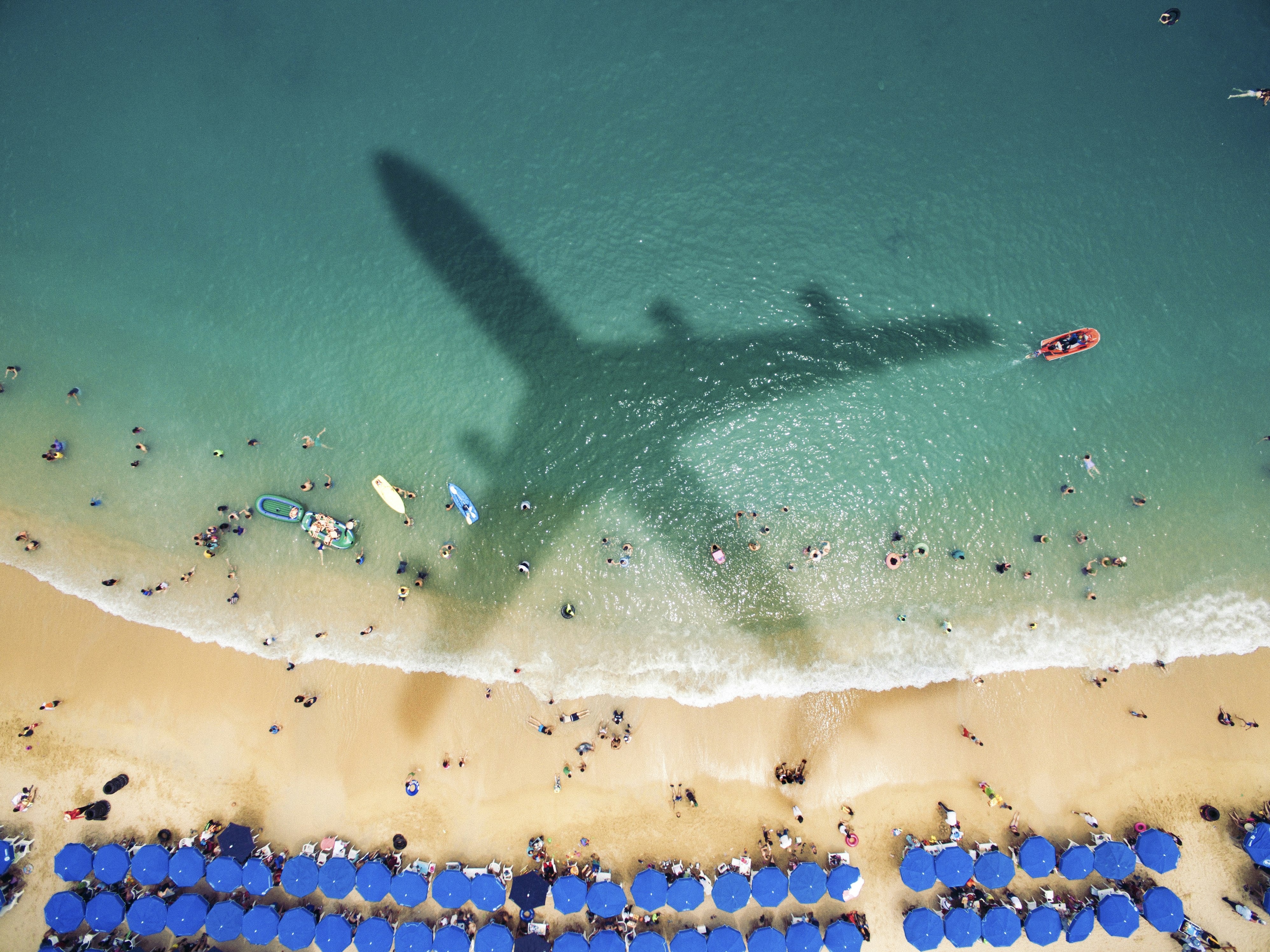 The shadow of a large jet plane falls over a beach with picturesque white sand, deep turquoise waters, and two long, waving rows of blue beach umbrellas. In the white surf break are the tiny dots of swimmers and beachgoers, as well as a red boat and a couple inflatable beach toys and two paddle boards.