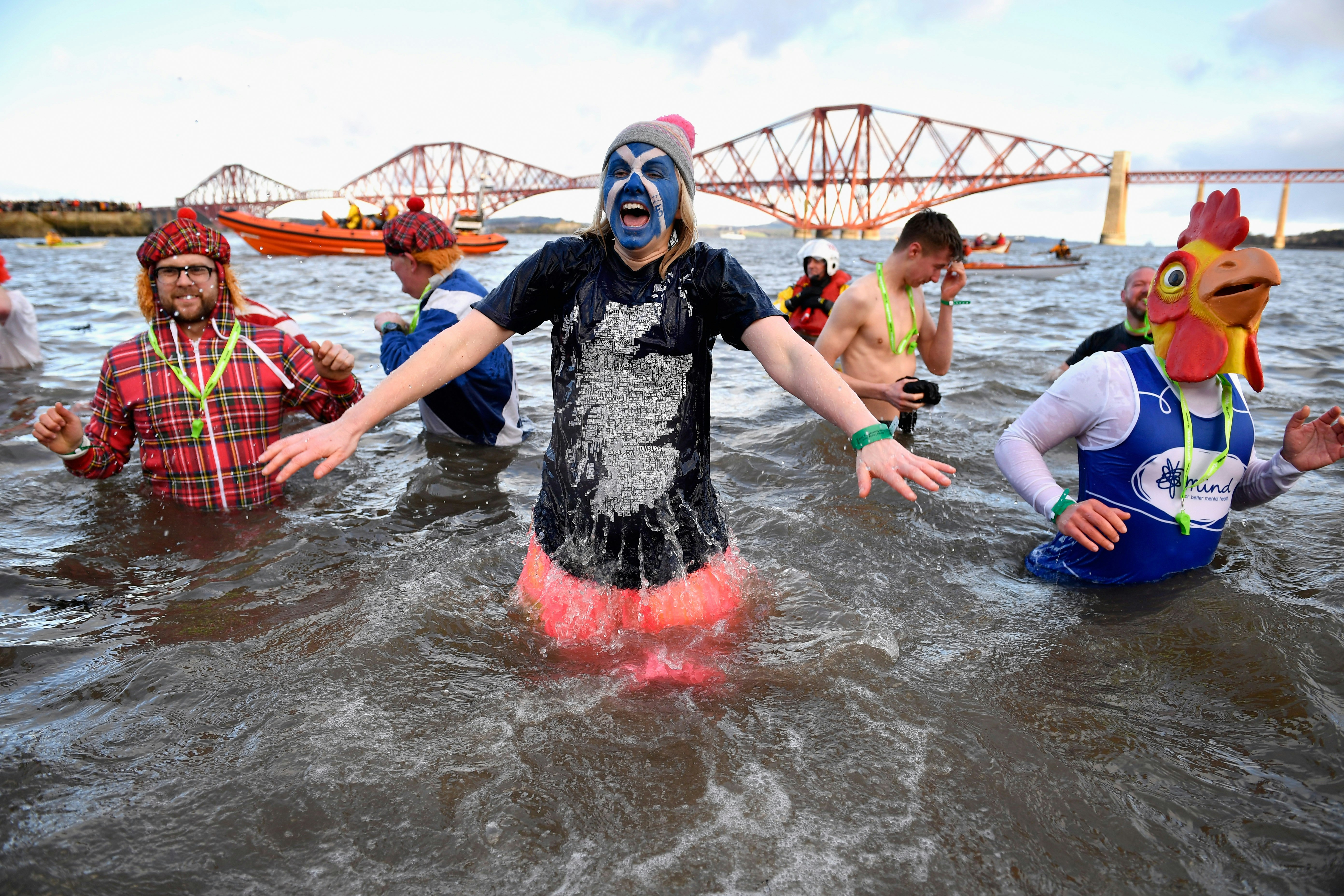 A group of people in fancy dress, one with the Saltire flag painted on their face, are wading in the River Forth and bracing themselves against the cold water. The iconic Forth Bridge can be seen in the background.