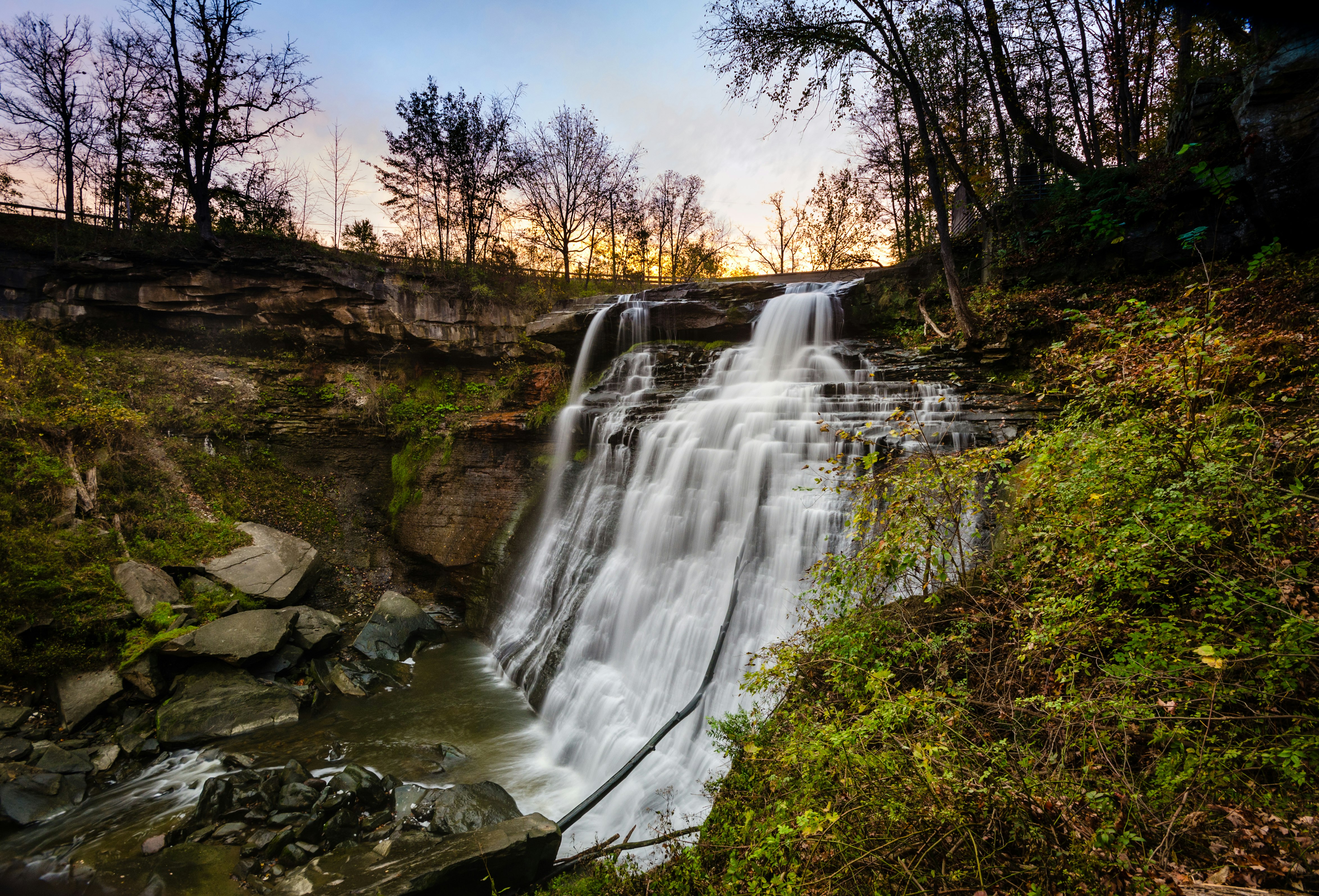 White water spills over narrow stair-stepped rocks that form Brandywine falls in  in Cuyahoga National Park. All around is lush foliage from surrounding shrubs, while on the top of the falls are the black outlines of larger trees against a faded sunset