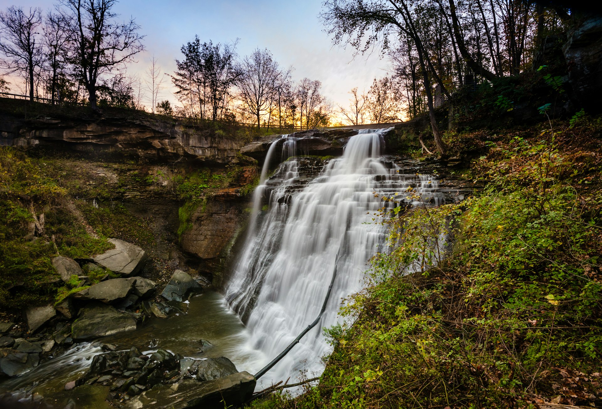 White water spills over narrow stair-stepped rocks that form Brandywine falls in  in Cuyahoga National Park. All around is lush foliage from surrounding shrubs, while on the top of the falls are the black outlines of larger trees against a faded sunset