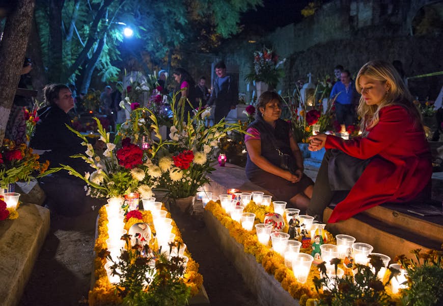 Two women sit beside a pair of candle-lit graves at night in Oaxaca, Mexico. In the background other people eye other decorated tombstones. 