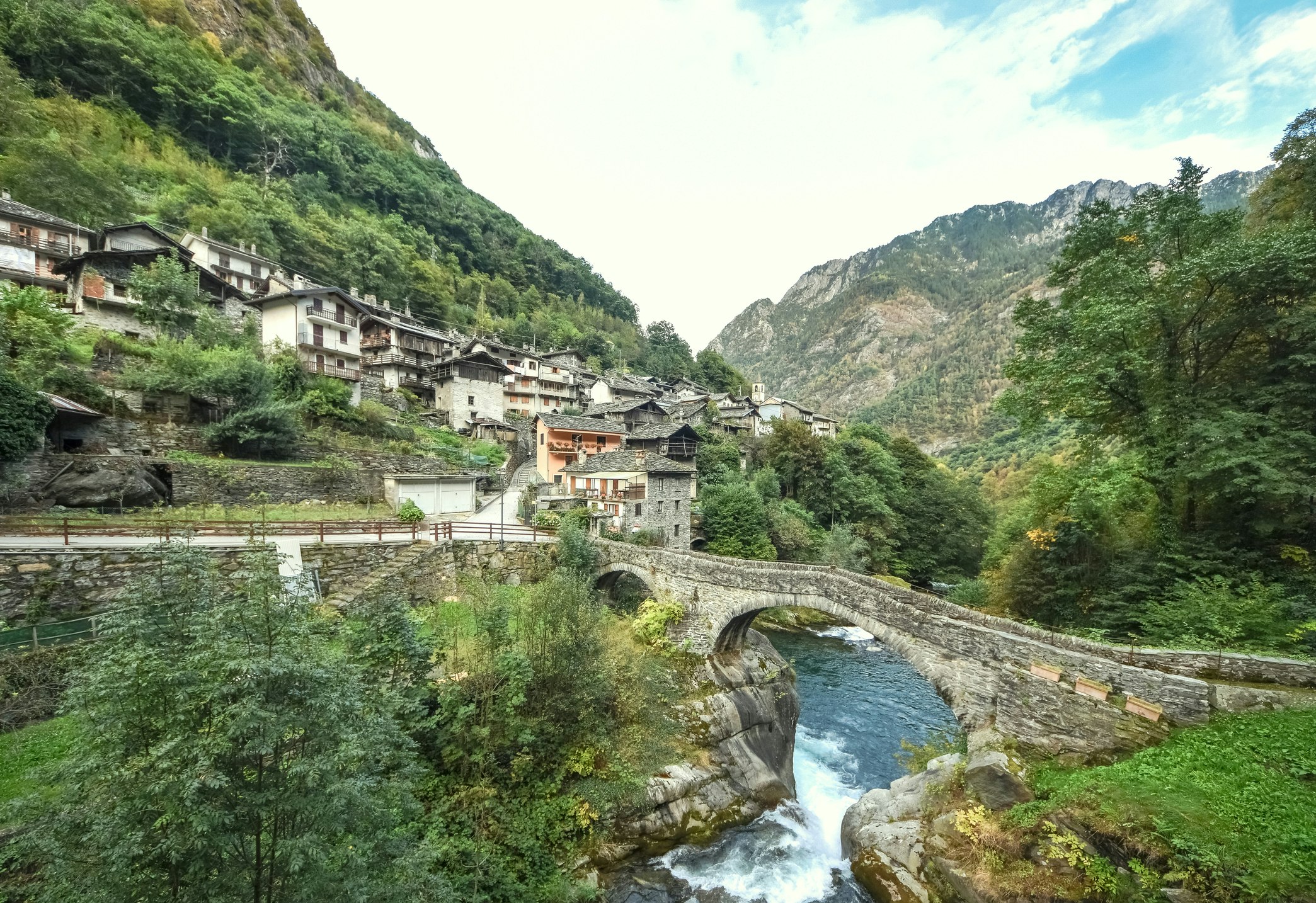 Alpine village with green mountains, wooden chalets and an old medieval bridge across the river