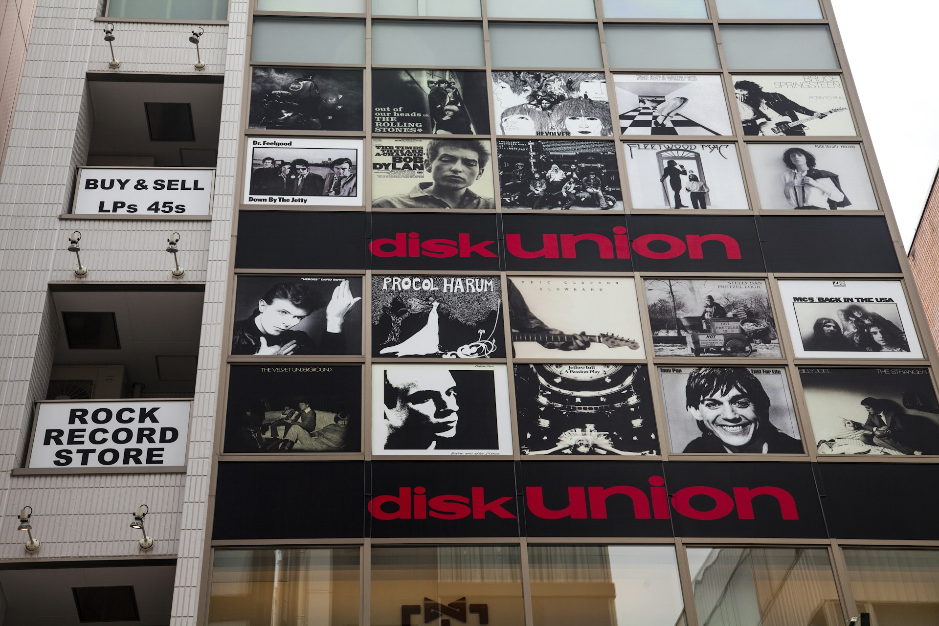 The facade of the disk union building in Tokyo's Shinjuku neighborhood puts classic album art in black and white in each window pane, so the building looks like a record bin. Between every two rows of picture-filled windows are long black signs that say "disk union" in a red san serif font.