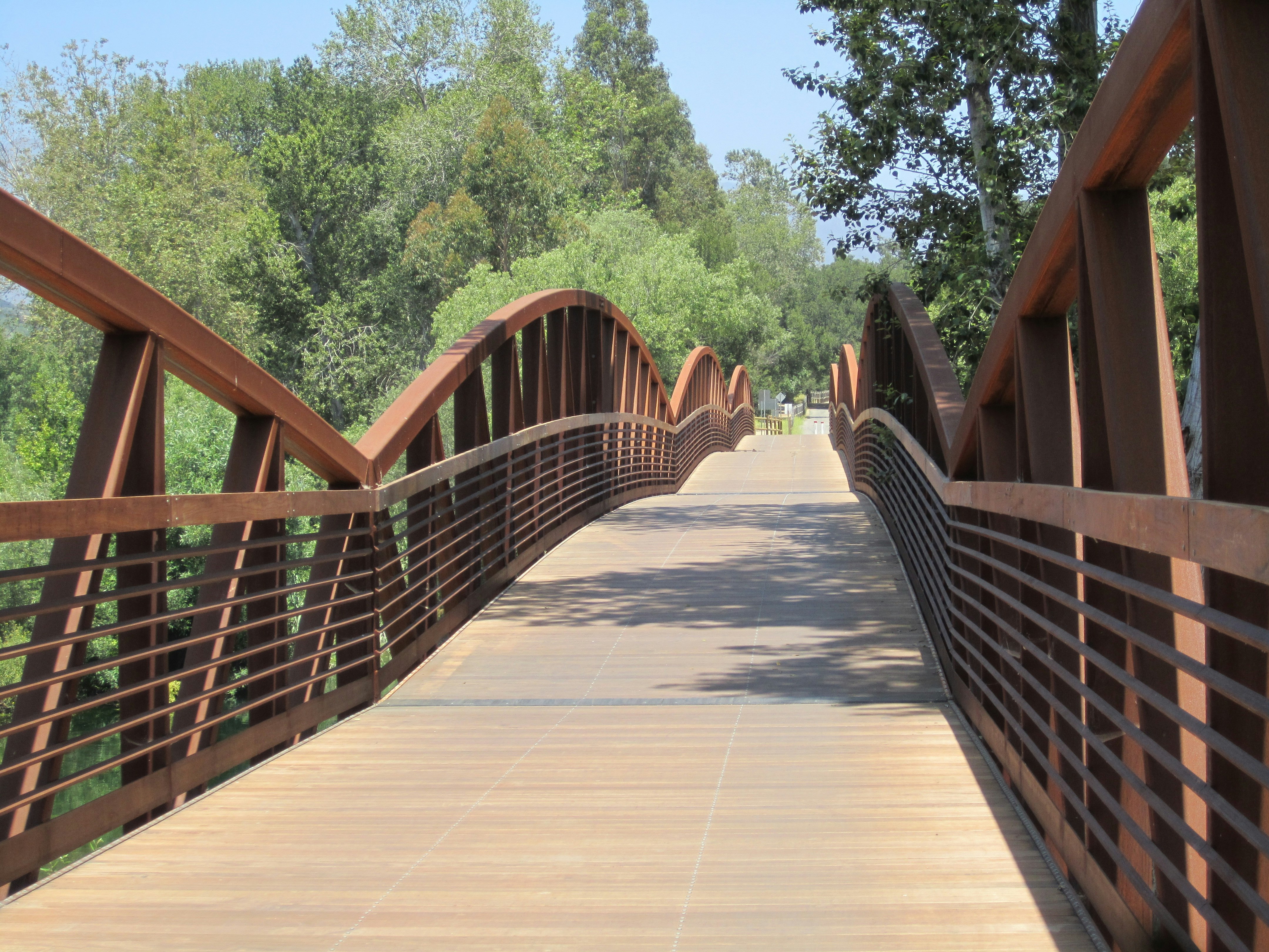 An orangey brown metal bridge made up of several horizontal slats under an arching, undulating portion that looks like the mountains in the area is filled in with a light brown wooden deck. The bridge itself undulates slightly and is surrounded by trees. It's part of the Ventura Trail rail to trail conversion to Ojai.