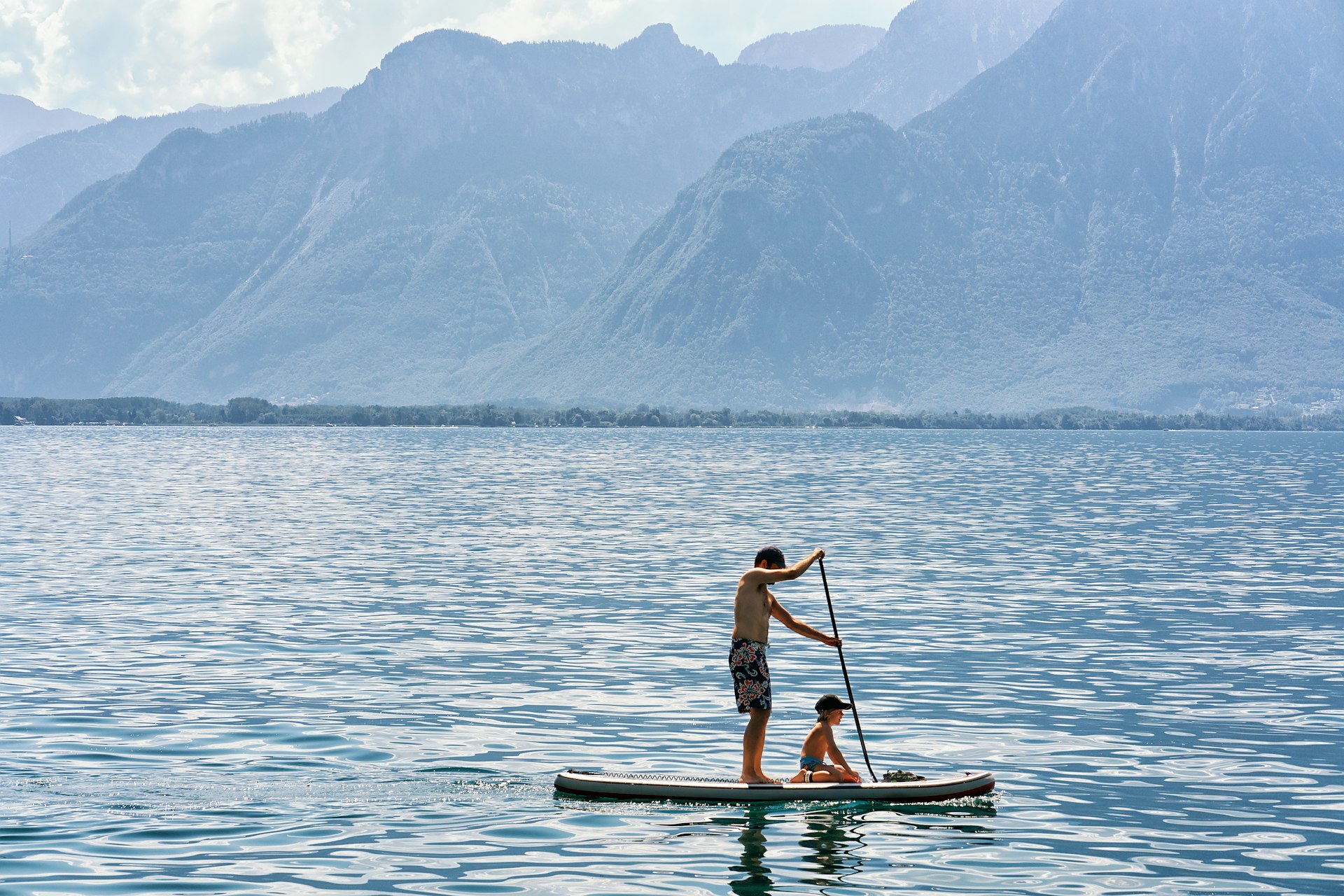A young man  paddle boards along placid denim-blue waters almost the same color as the steep mountains behind the lake. He wears blue and green swim trunks and a toddler in a baseball cap sits on the front of the paddle board.