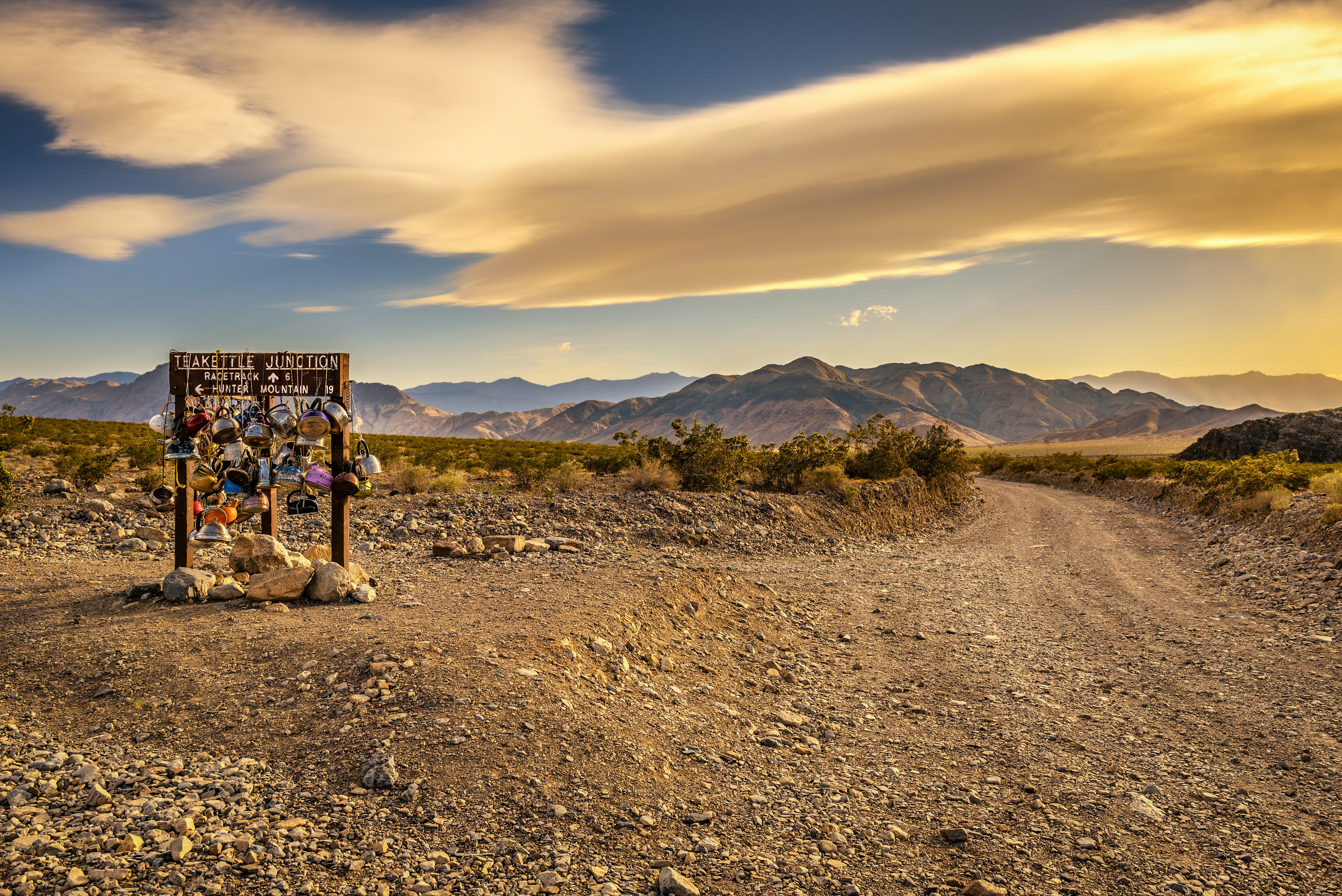 The golden expanse of Death Valley's rocky, sandy flats surround a worn wooden sign reading "Teakettle Junction" on which are hung numerous vintage kettle in various muted colors. A flat road winds off into to the distance towards mountains tinted blue and purple.