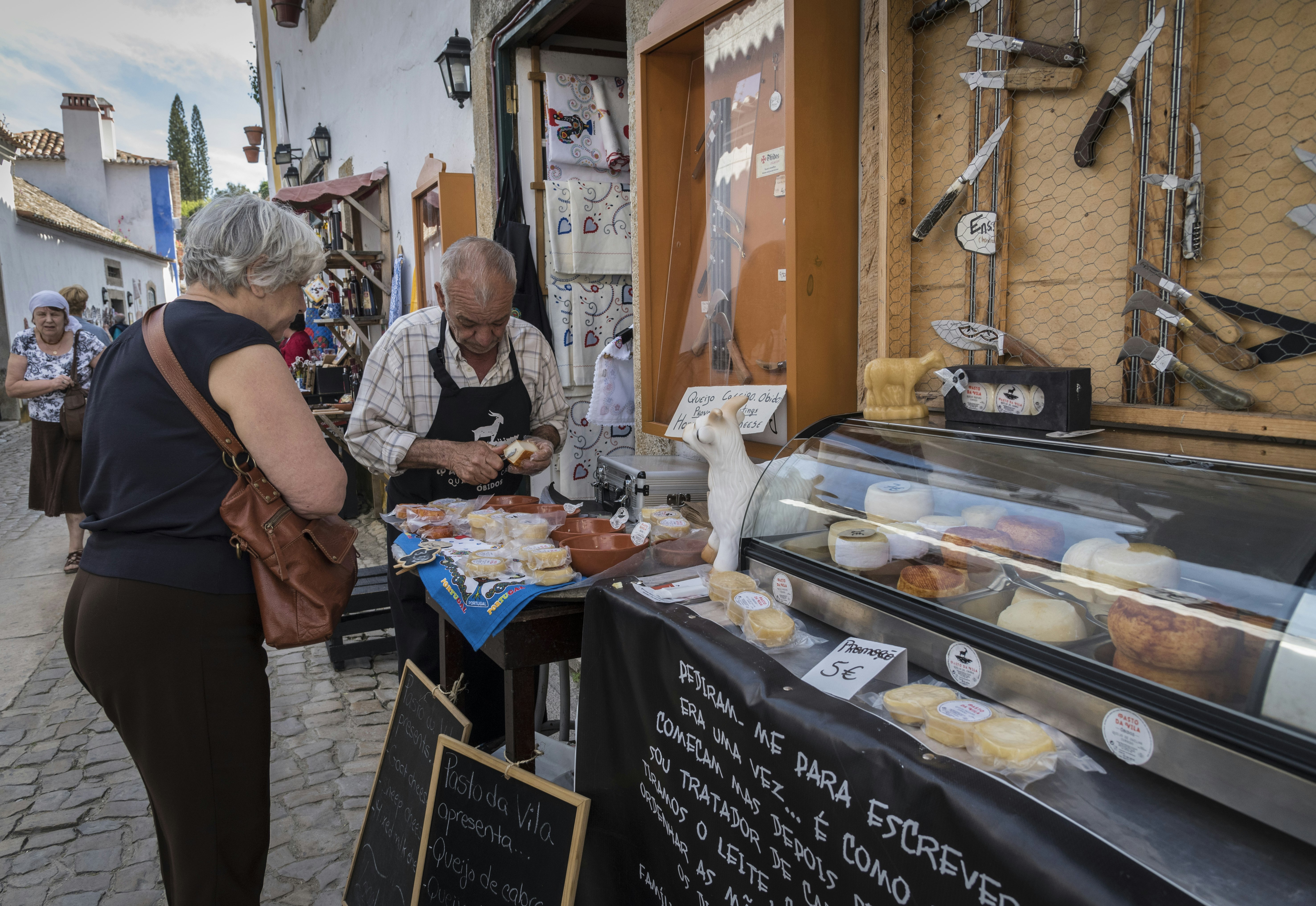 A shopkeeper is slicing a piece of cheese for a customer to taste from a stall outside a shop. Many different types of cheese are on display and an array of culinary knives adorn the wall above a glass cabinet.