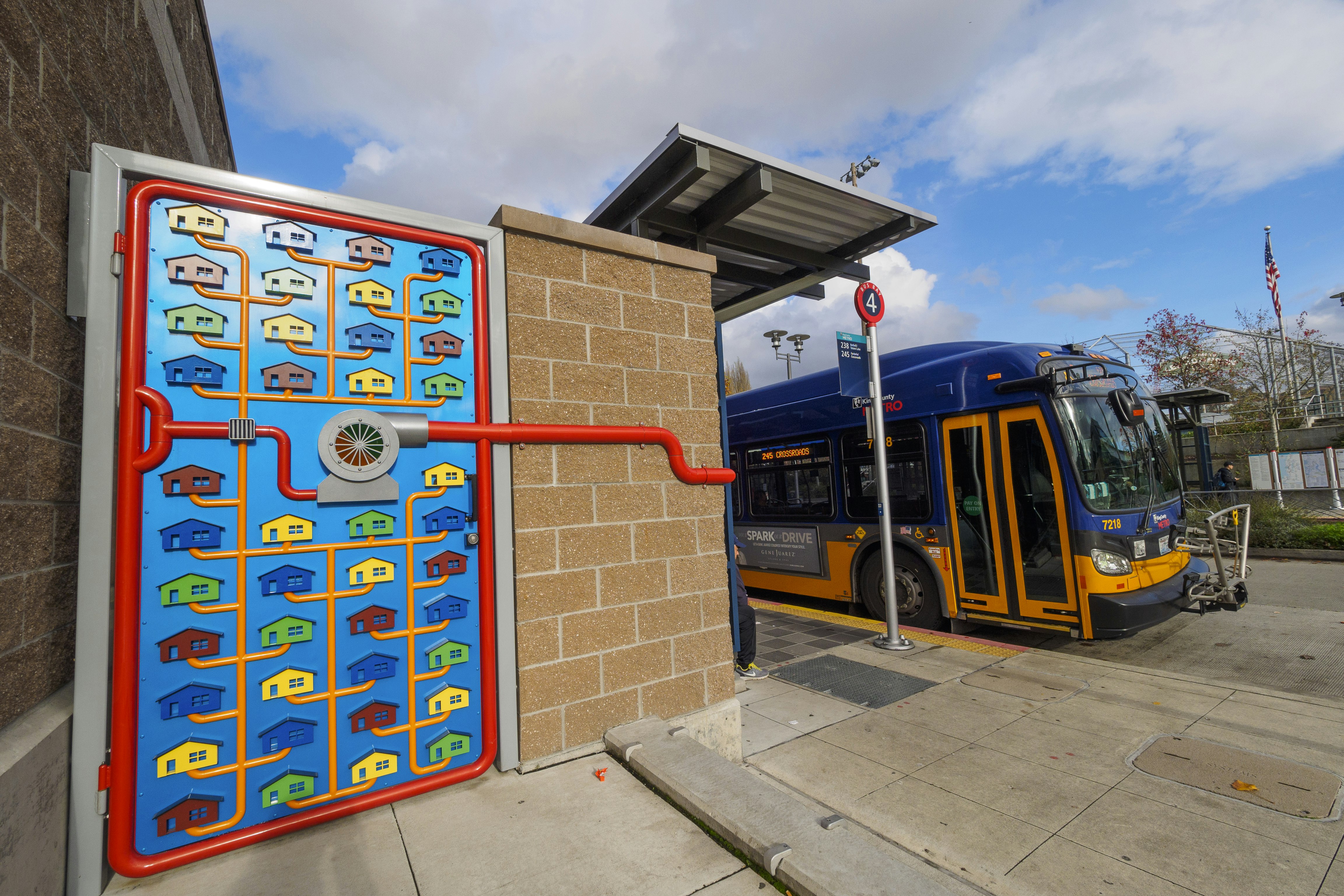 A gate made out of red metal piping and colorful metal depicts four columns with eleven rows of styled houses in primary colors, all connected by orange plumbing. This piece of public art in Kirkland, Washington adorns the Pump Station Gates. A bus stop for the number 4 line sits in the background, with a blue and orange bus stopped by a beige sidewalk