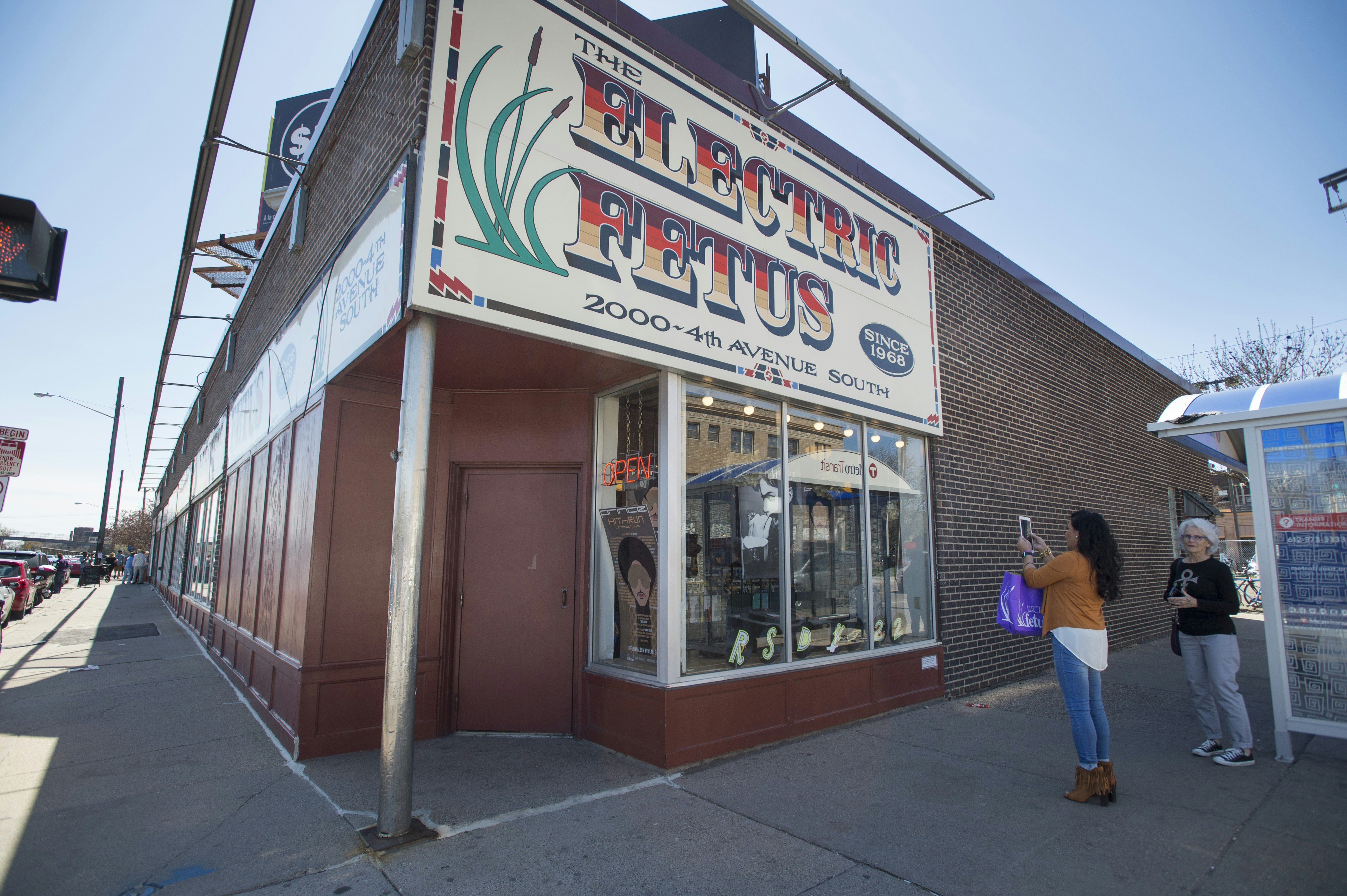 A woman in blue skinny jeans, brown booties, a white hi-lo top, and camel-colored sweater snaps a photo on her phone of the Electric Fetus building, a brick storefront with a large purple, orange, and tan 70s-style font and an illustration of cat tails with waving leaves.