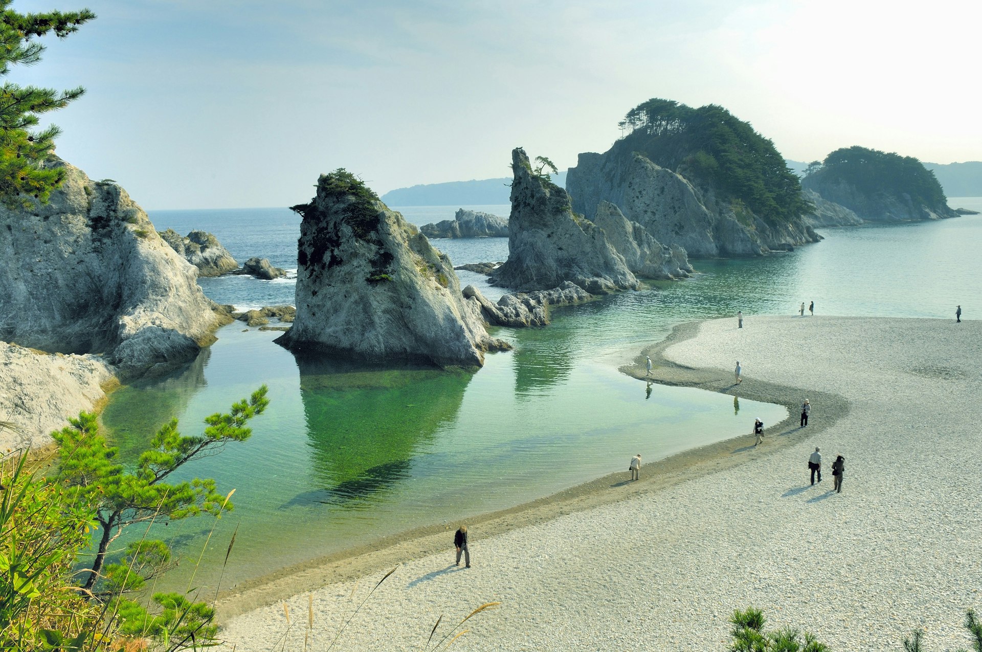 A selection of jagged rocks, the Jodogahama volcanic rock formations, poke out from the tranquil blue sea waters of northern Japan. The rocks are peppered with greenery, and a small crowd of people view and photograph them from the small strip of beach in the foreground.