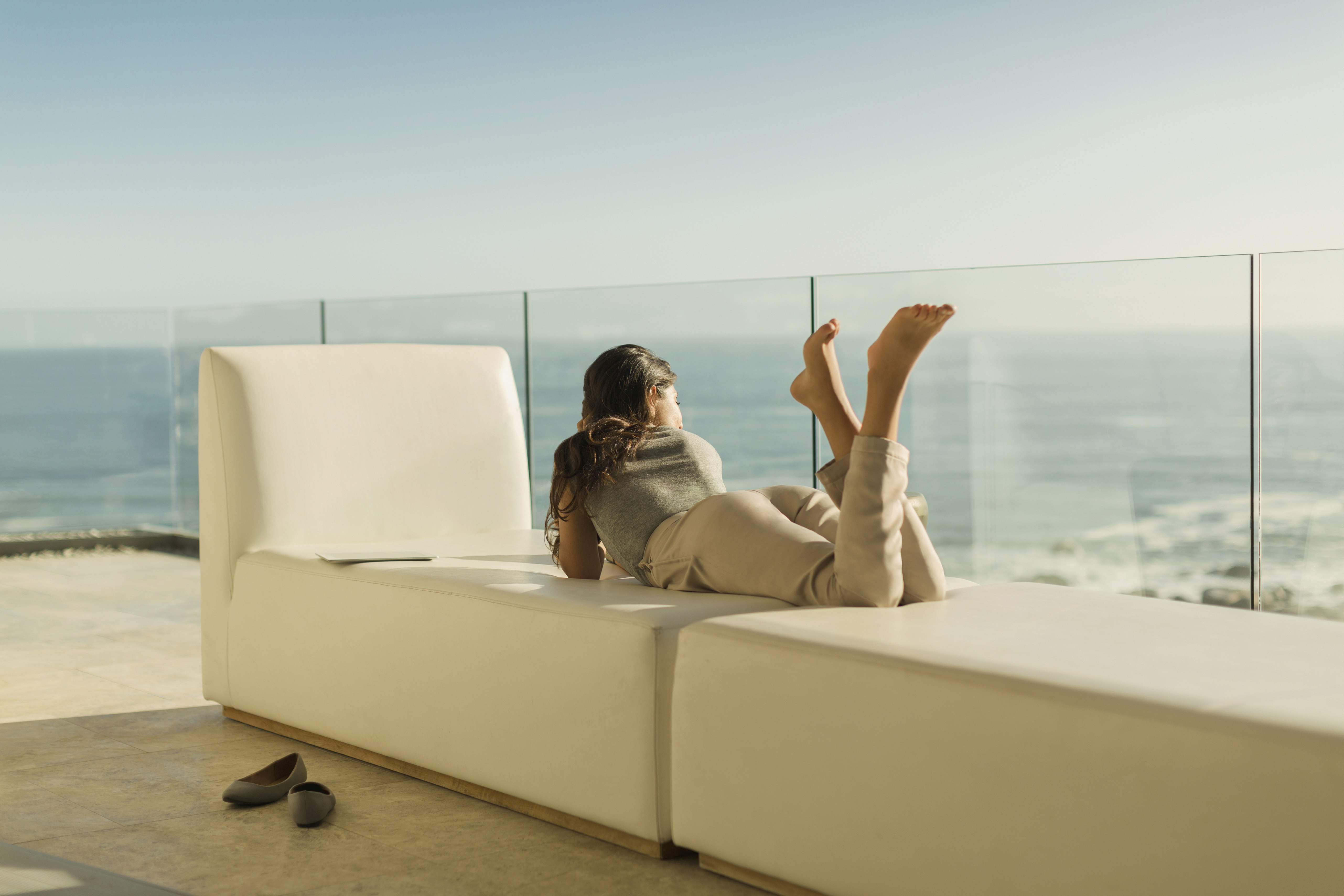 A woman lying on a cream-coloured sofa, her shoes on the floor; the setting is a glass-sided balcony on the edge of the coast, with the ocean in full view.