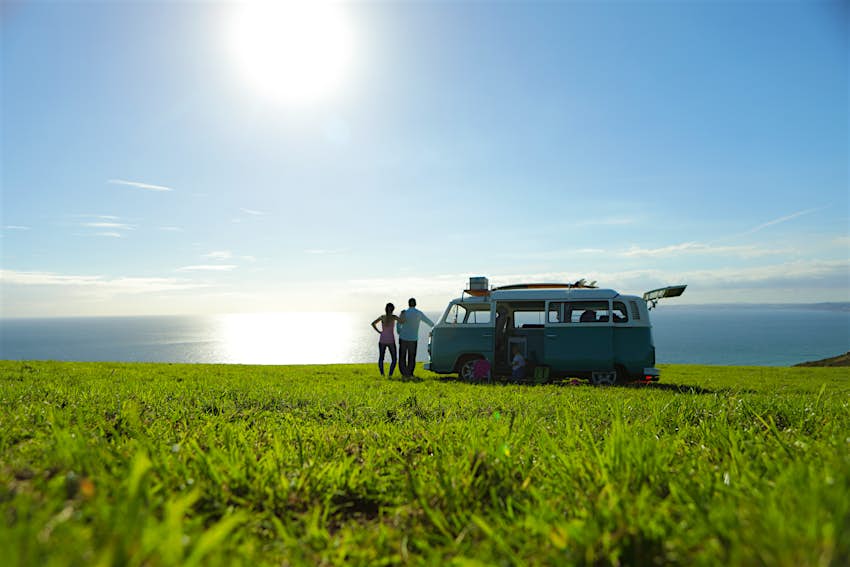 VW camper van in a field Two people stand next to a classic VW camper van parked in the middle of a grassy field. The sea is visible in the distance.