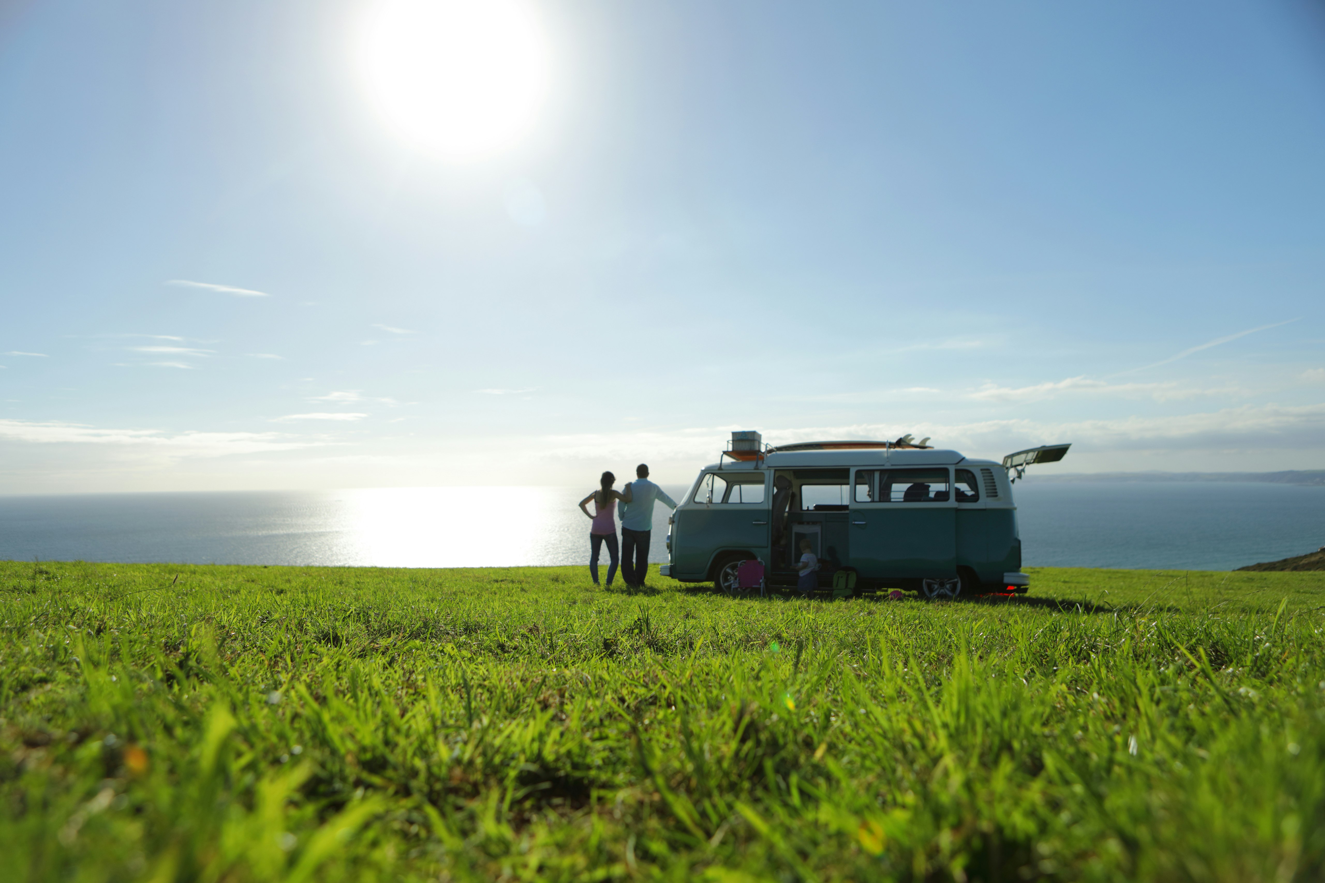 Two people stand next to a classic VW camper van parked in the middle of a grassy field. The sea is visible in the distance.