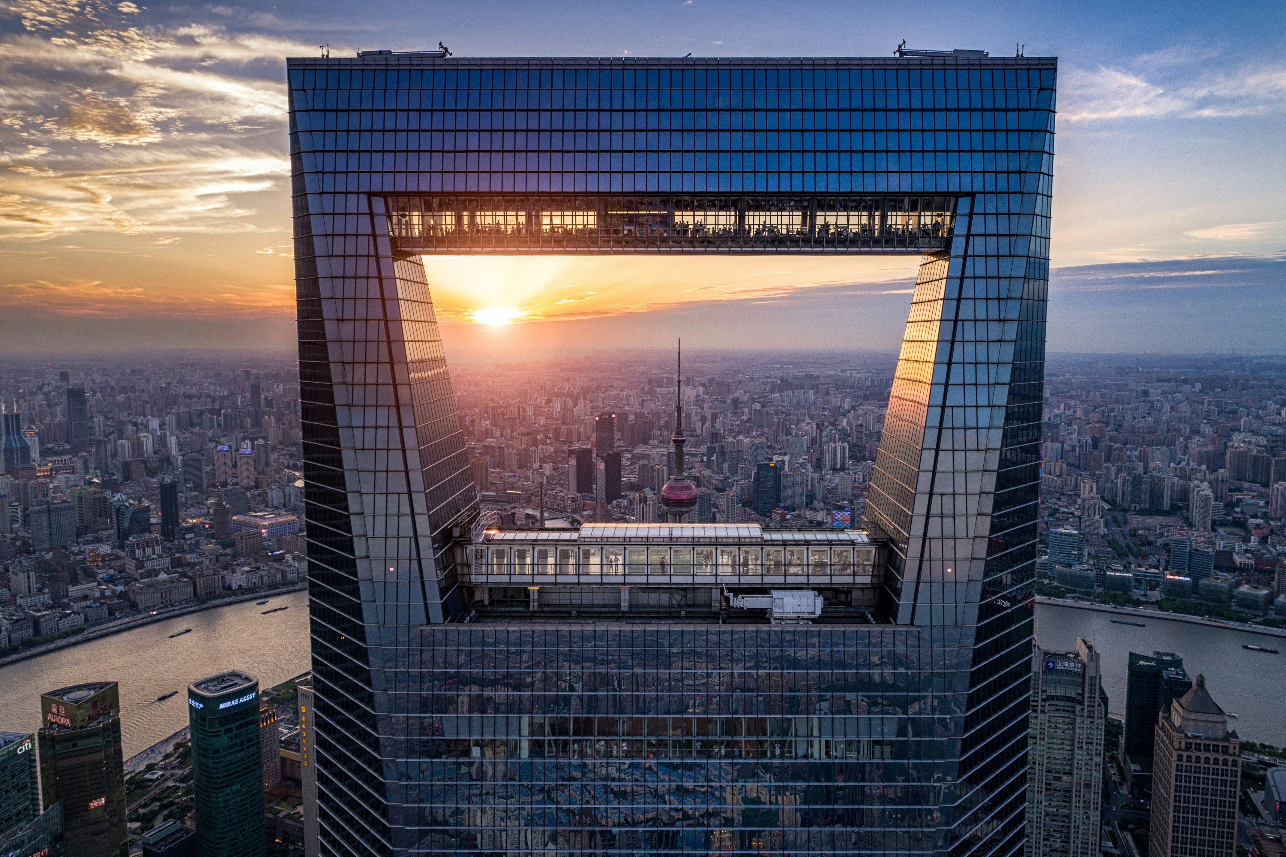 A view of the Shanghai World Financial Centre, a skyscraper with a square 'window' through the centre of it, which gives it the nickname of the bottle opener. Beyond the skyscraper the river and buildings of Shanghai are visible.