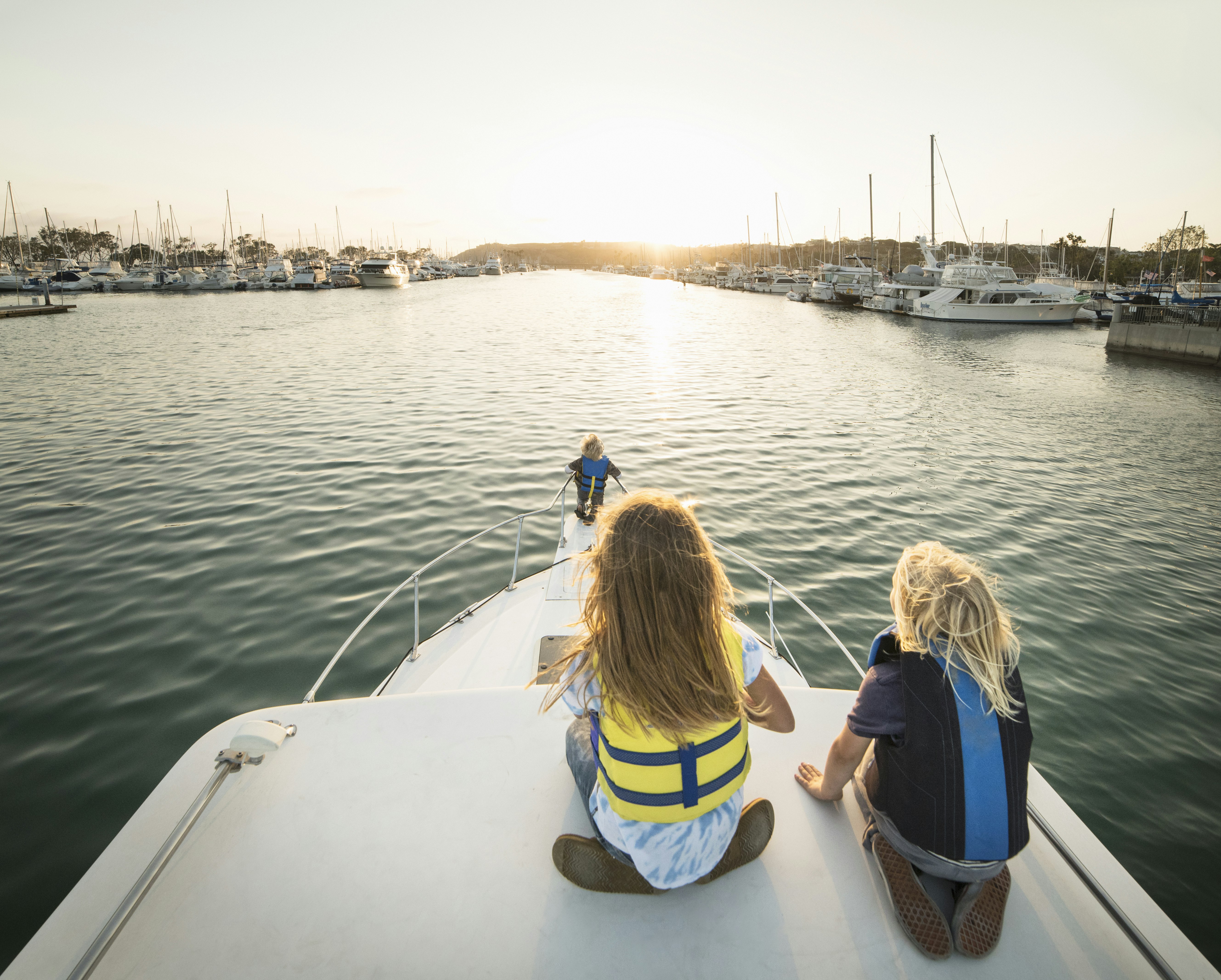 Three kids sit on the front of a boat as it cruises through the harbor