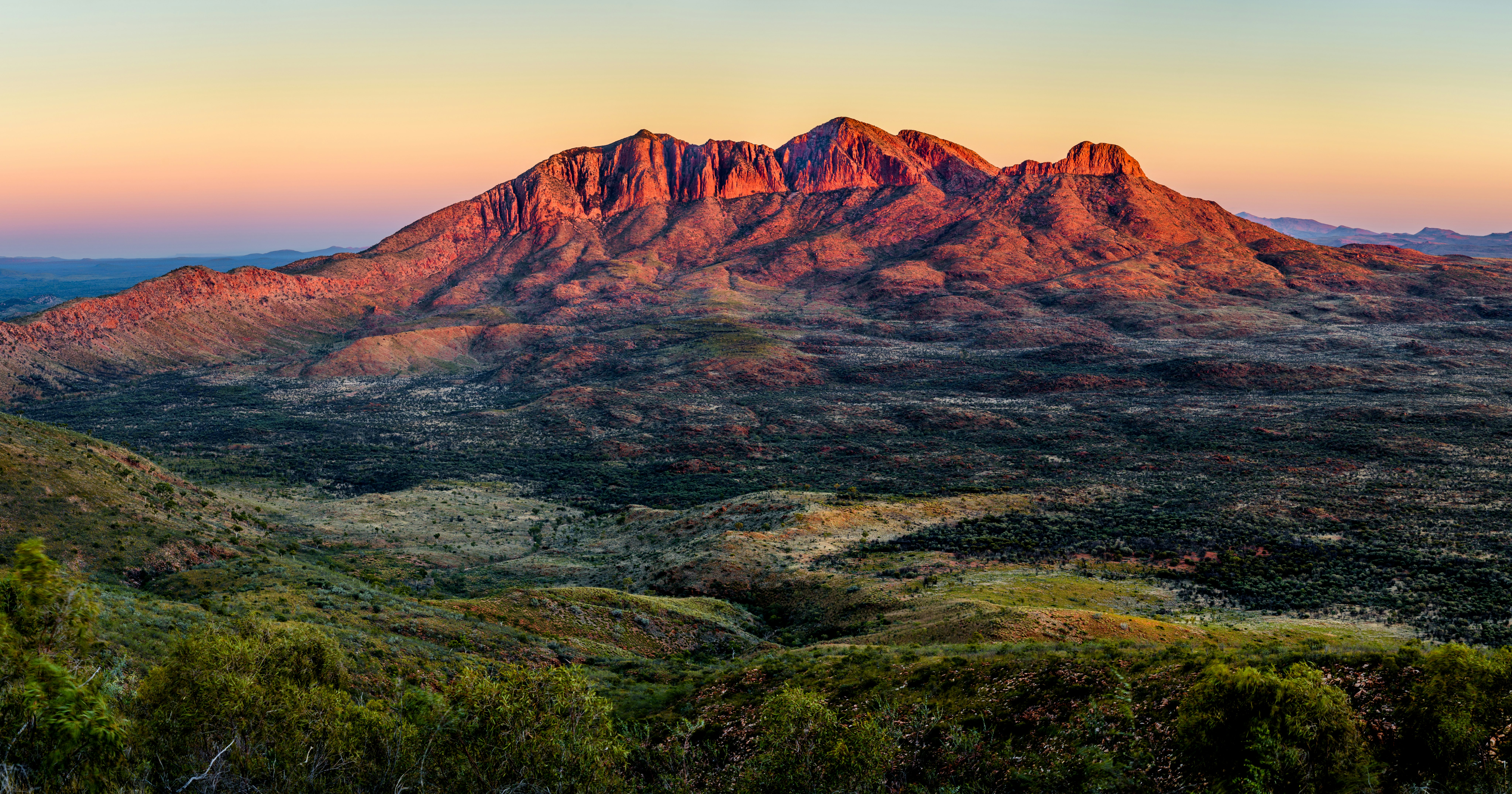 The deep red, orange, and purple alpenglow of Mt. Sonder in the West Macdonnel Range of Australia gleams at sunset over a variegated valley floor