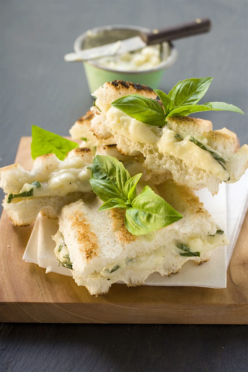A stack of toasted tramezzini, filled with melted mozzarella and chives, sitting on white paper napkins a wooden chopping board. Sprigs of basil have been artfully placed as decoration.